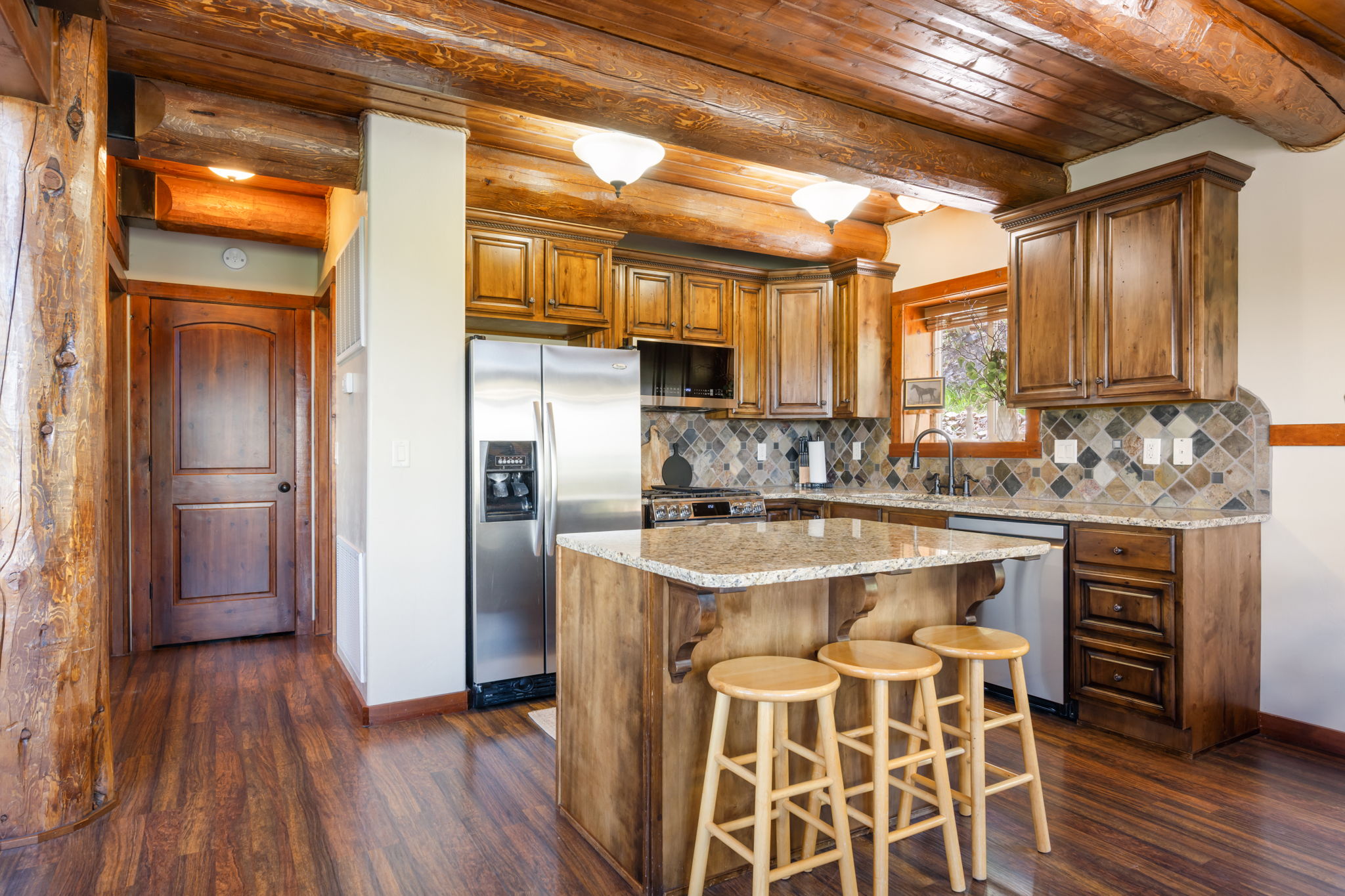 Open-concept kitchen featuring wood-finish flooring, exposed log beams, granite countertops, wood cabinetry, and a tile backsplash with diamond accents