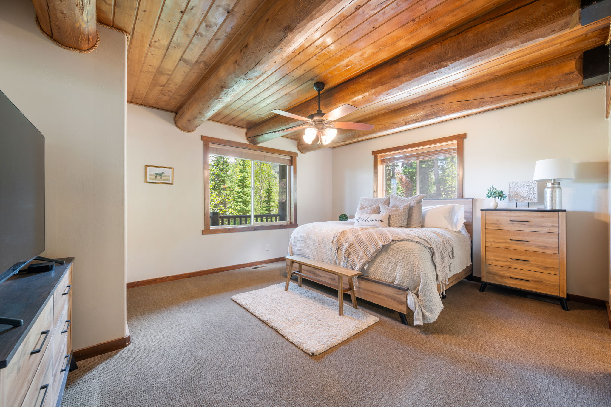 Exposed log beam ceiling with a ceiling fan, neutral carpet flooring, and two large windows featuring wood trim