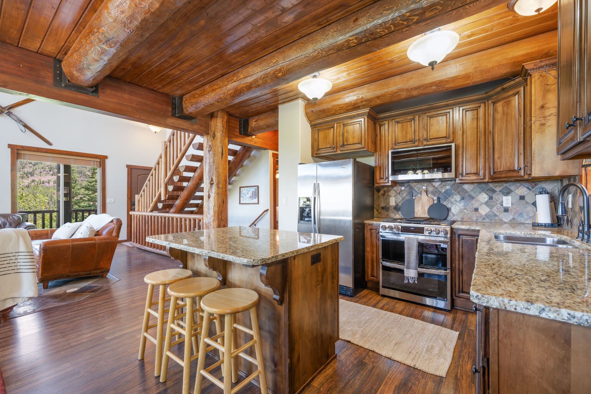 Kitchen and living area featuring exposed wood beams, wood-finish flooring, a kitchen island with stone-finish countertop, stainless steel appliances, and a wood staircase
