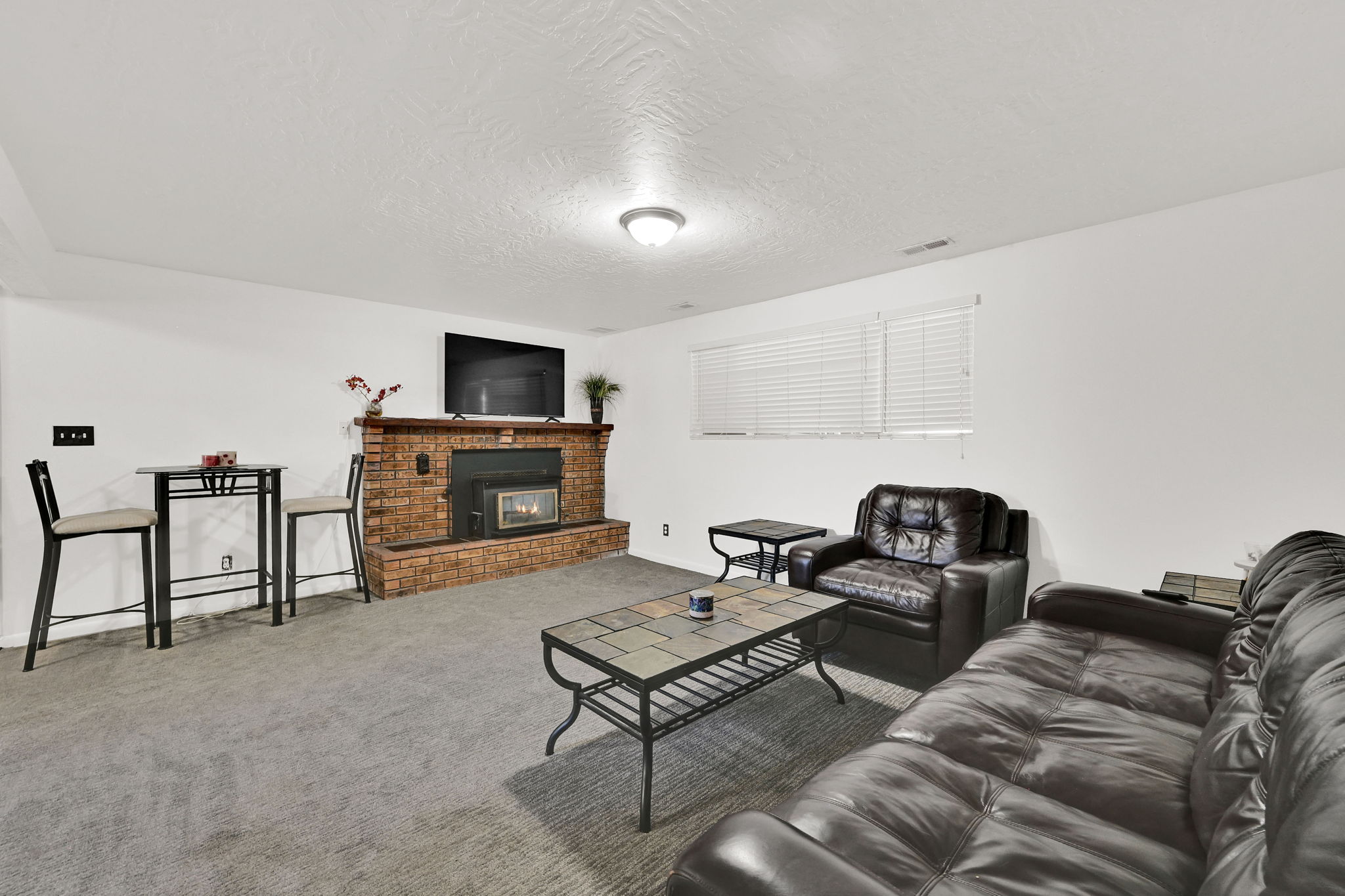 Living area featuring a brick fireplace with a raised hearth, light gray carpet flooring, white walls, and a window with horizontal blinds