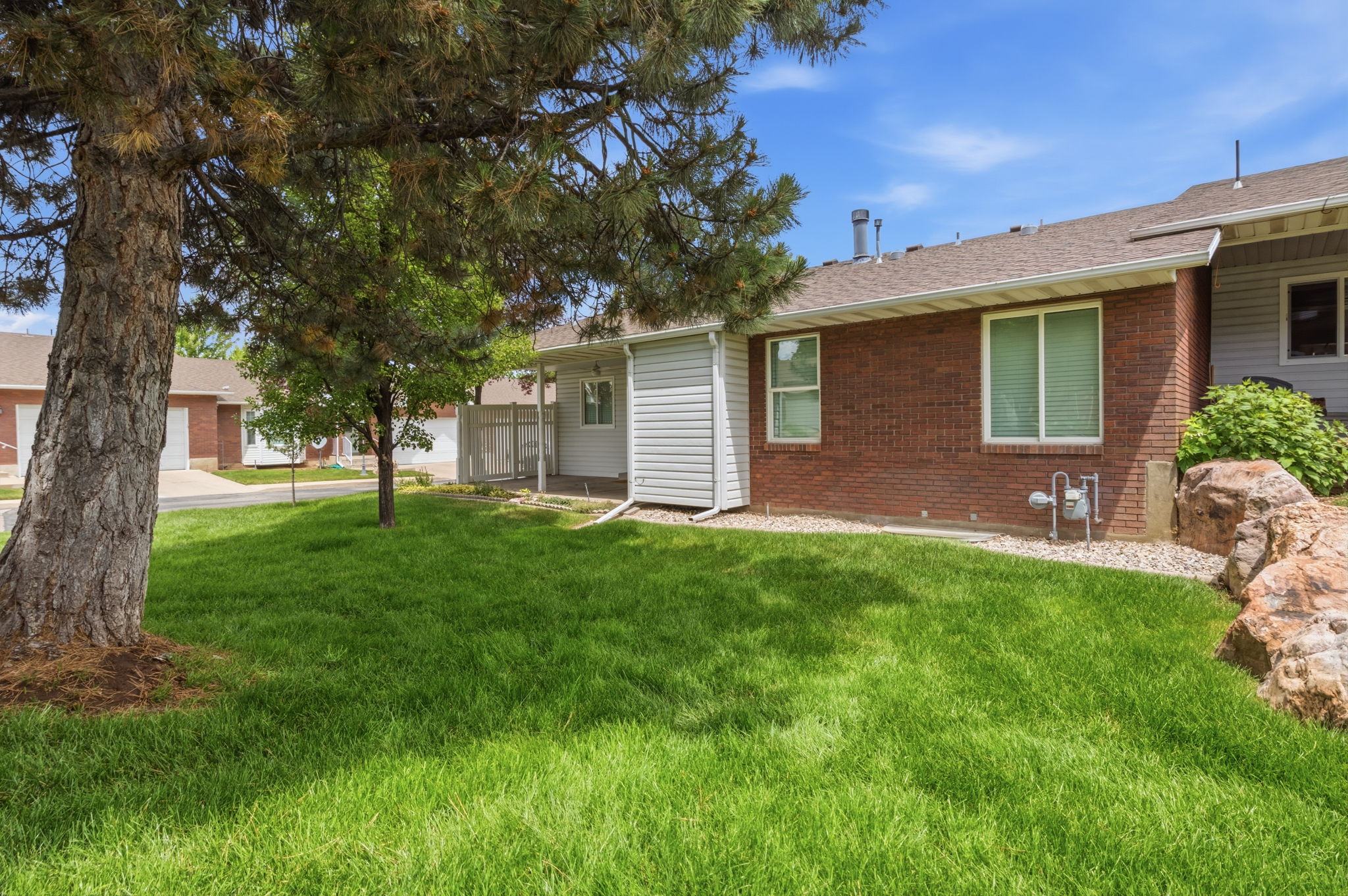 Green lawn area featuring a large mature tree, a red brick exterior, white siding, and a partial rock garden with natural boulders
