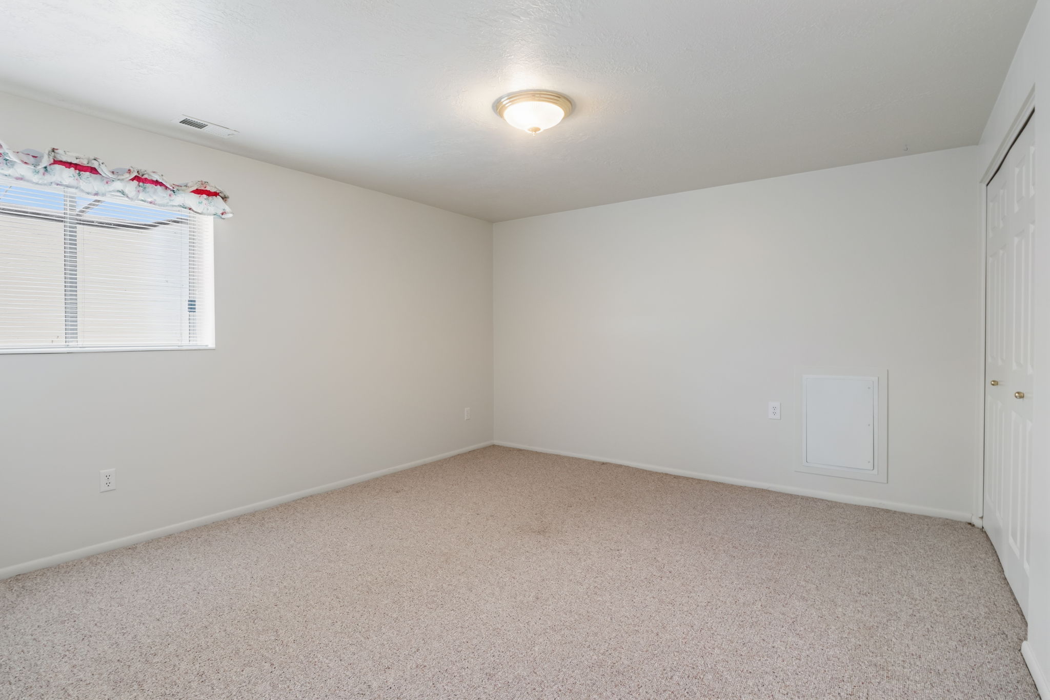 Spacious room featuring neutral carpet, a single window with white blinds, a flush-mount ceiling light fixture, and a white bi-fold door