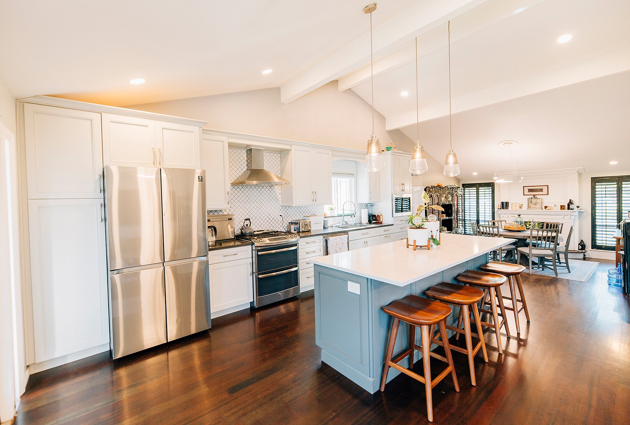 Open-concept kitchen featuring a spacious island with seating, stainless steel appliances, white shaker-style cabinetry, dark wood-finish flooring, and vaulted ceilings with exposed beams