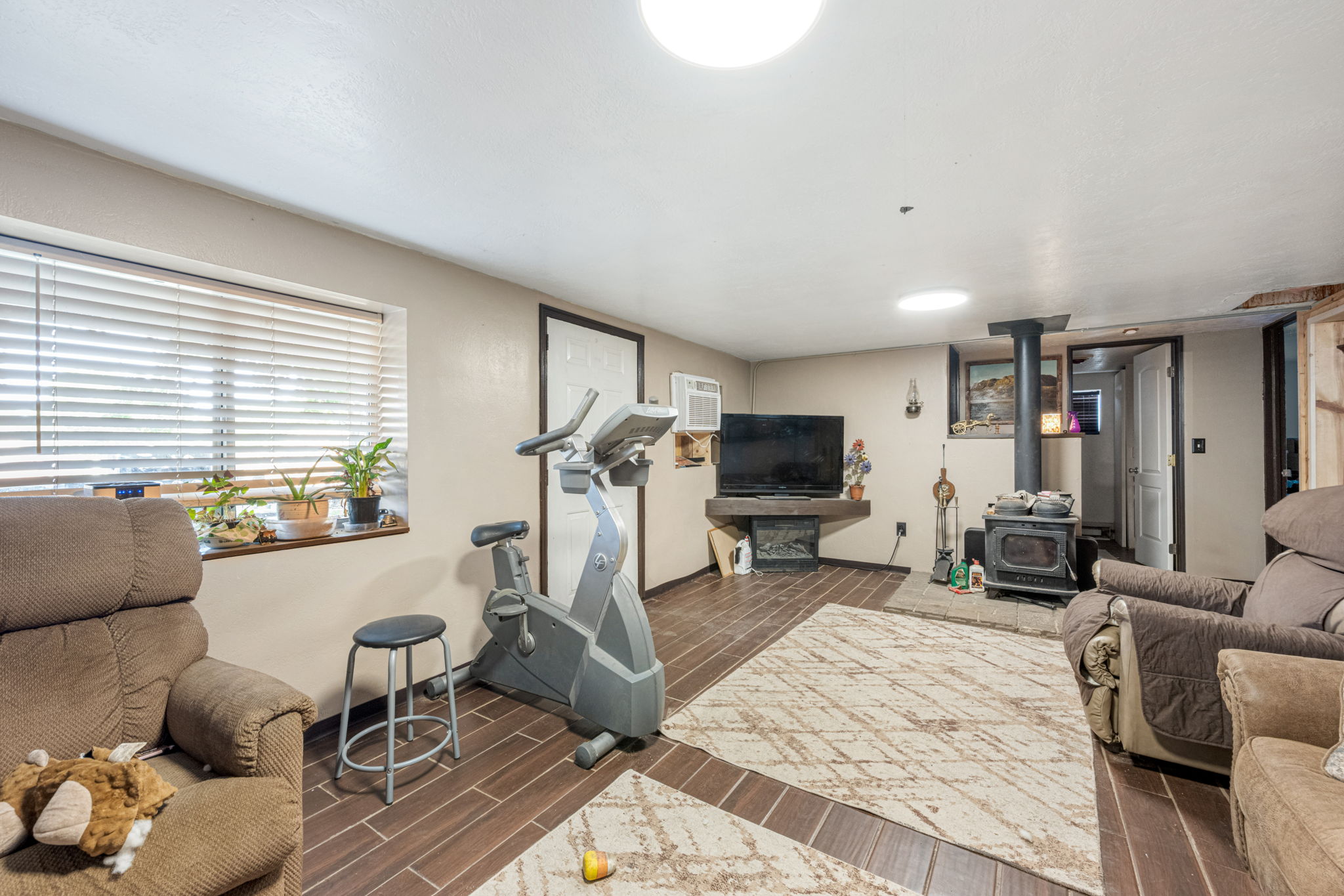 Spacious room featuring wood-finish tile flooring, a window with blinds, and a wood-burning stove