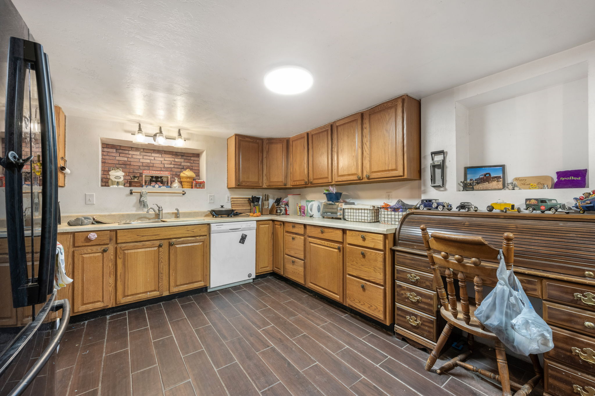Kitchen featuring wood-finish flooring, wood cabinetry with upper and lower storage, a built-in dishwasher, an integrated sink, and an exposed brick backsplash with overhead lighting