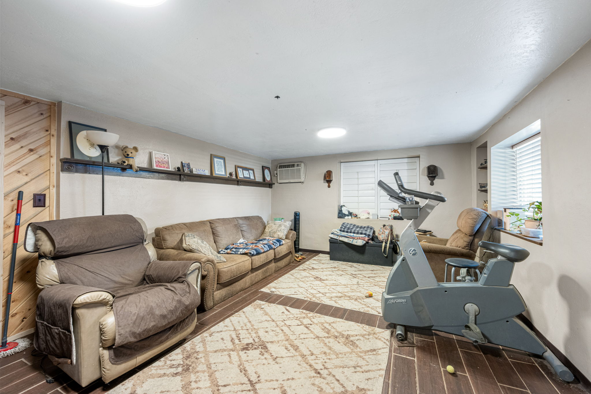 Spacious room featuring wood-finish flooring, light-toned walls, a built-in wall shelf, and two windows with horizontal blinds