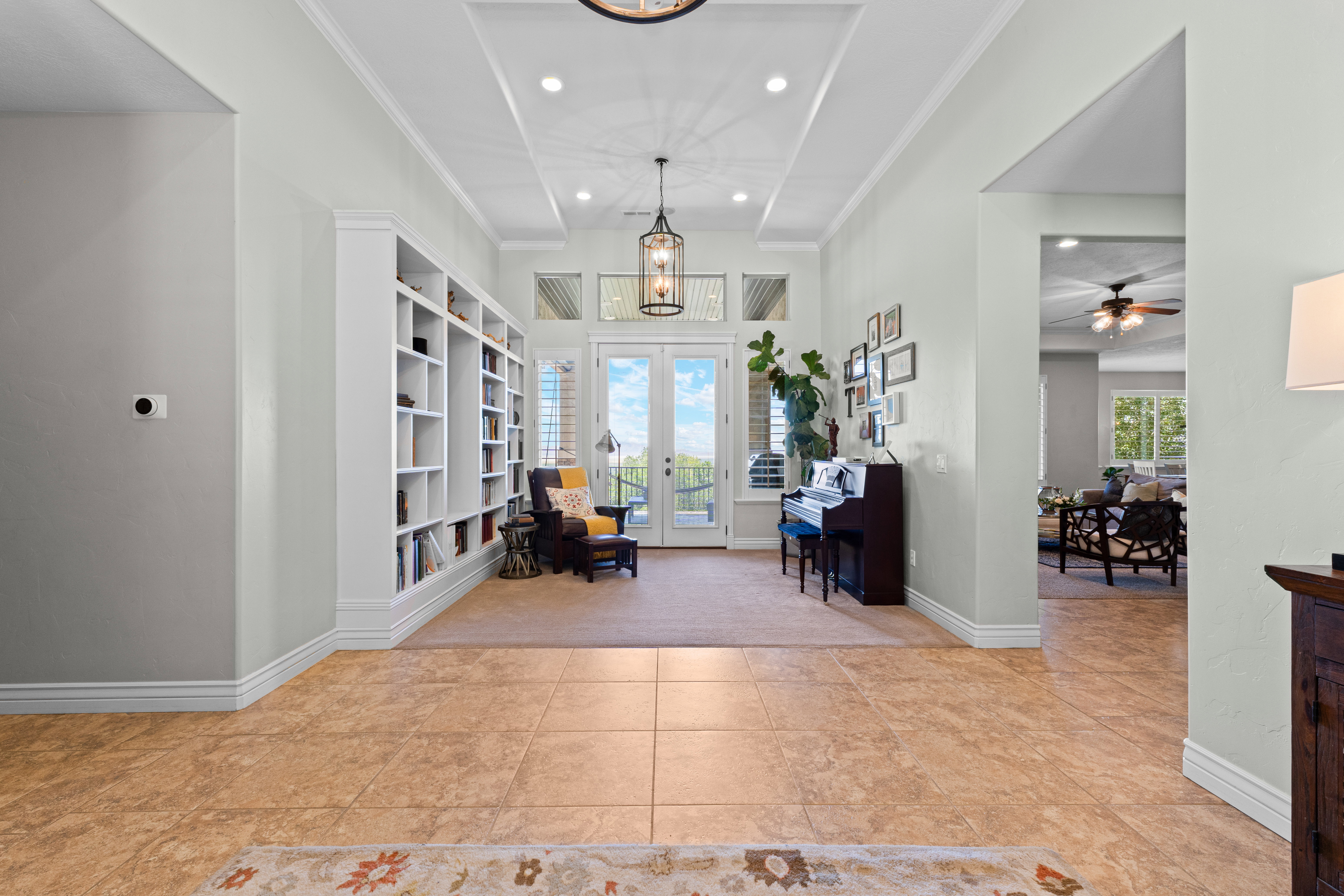 Grand entry hall featuring large format tile flooring, a coffered ceiling with recessed lighting, an extensive built-in bookcase, a double glass-panel door, and an overhead chandelier