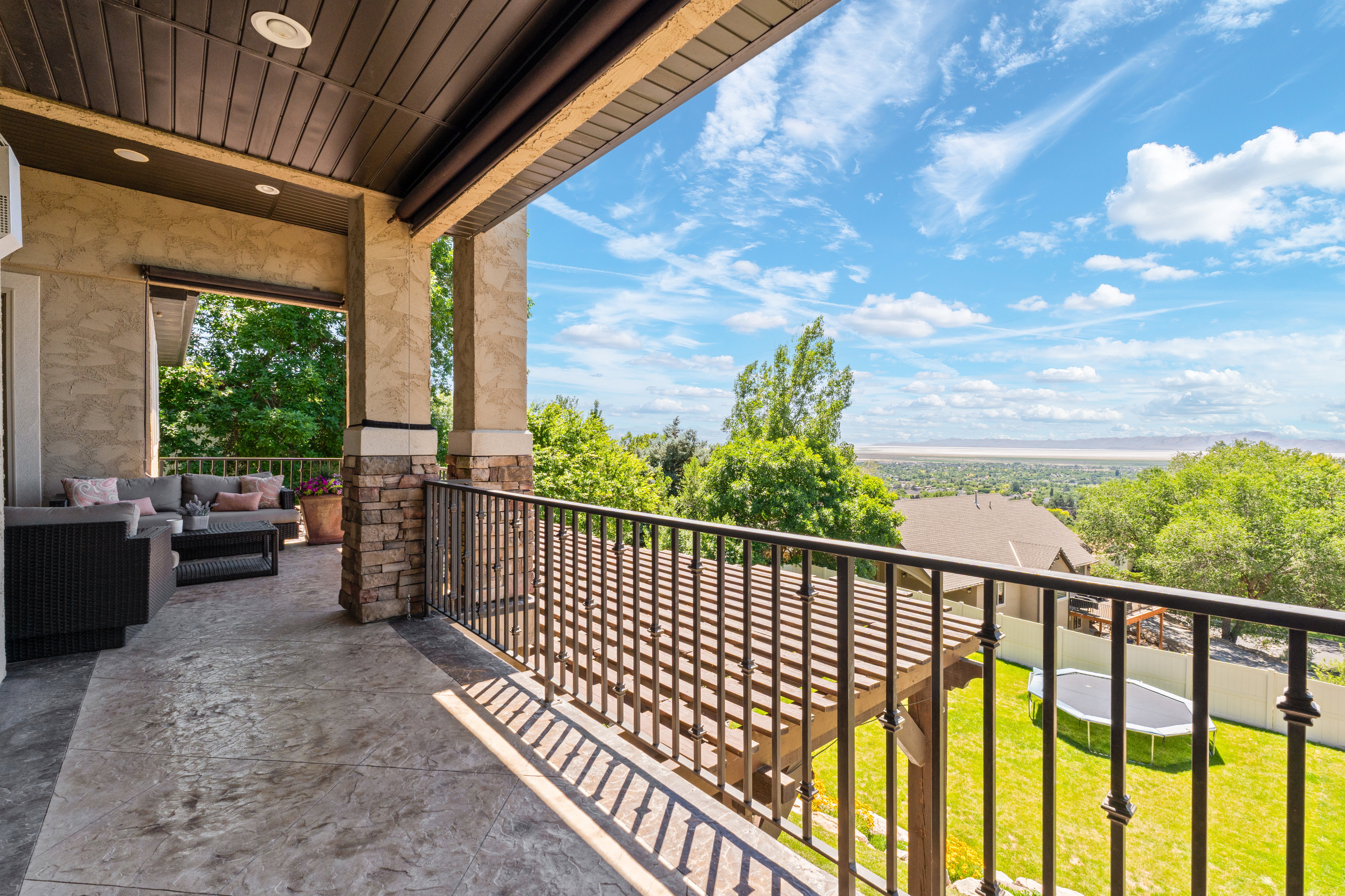 Expansive covered balcony featuring stamped concrete flooring, a detailed metal railing, and stone-clad columns