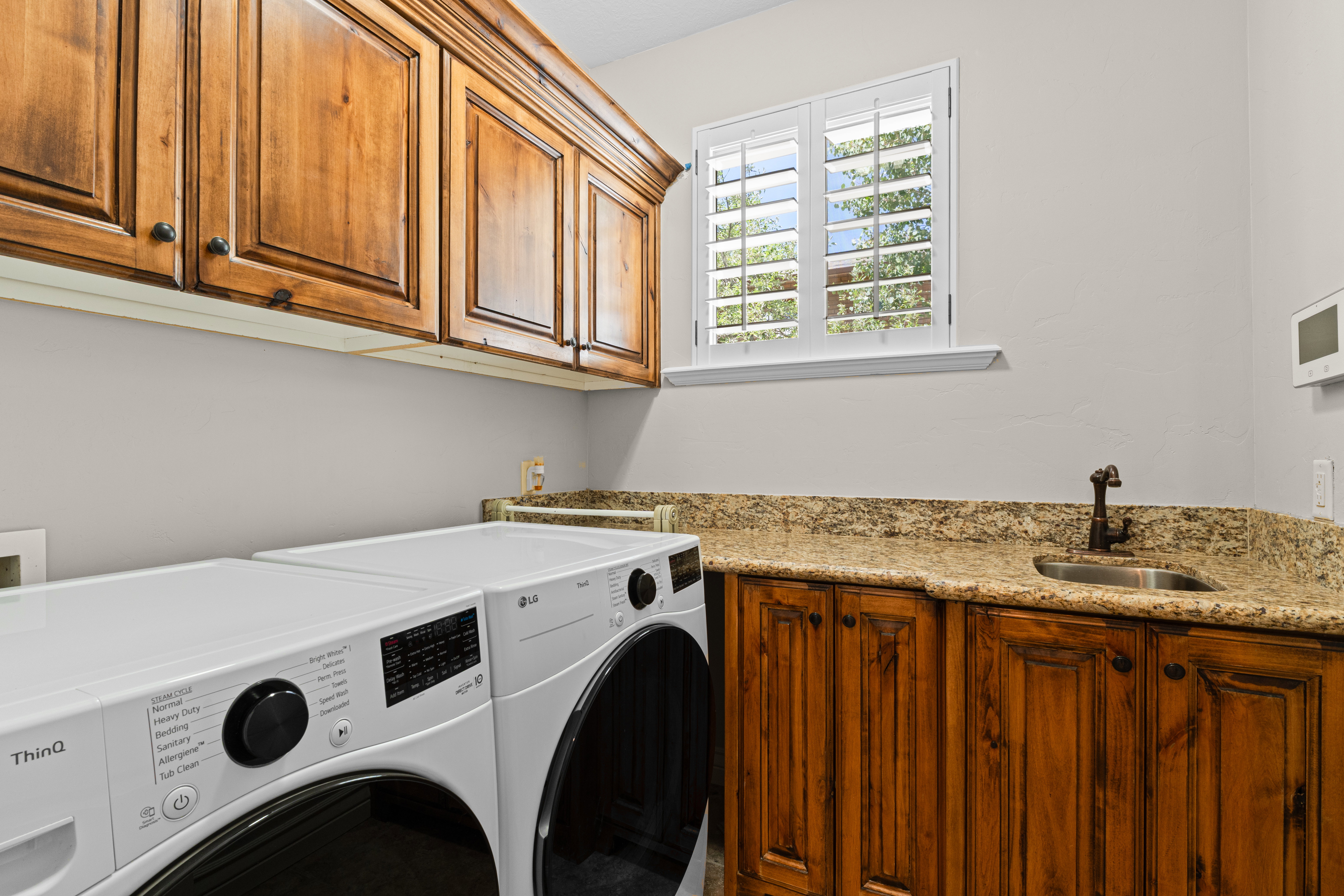 Utility room featuring wood cabinetry, granite-finish countertops, stainless steel sink with bronze-tone faucet, and plantation shutters