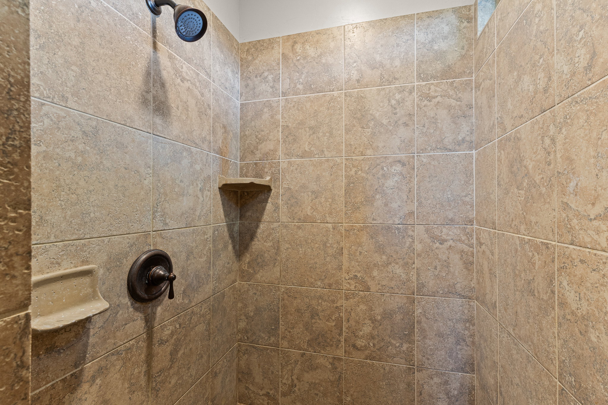 Tiled shower featuring a bronze showerhead, coordinating control handle, and built-in corner shelving