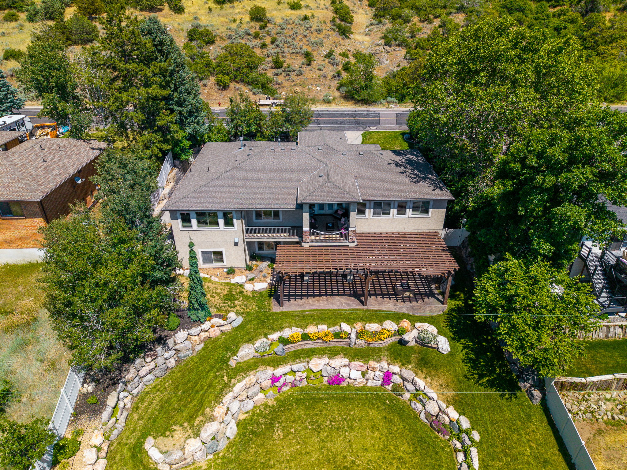 Two-story stucco property featuring a large backyard with a multi-level retaining wall, mature trees, and a wooden pergola covering a patio space