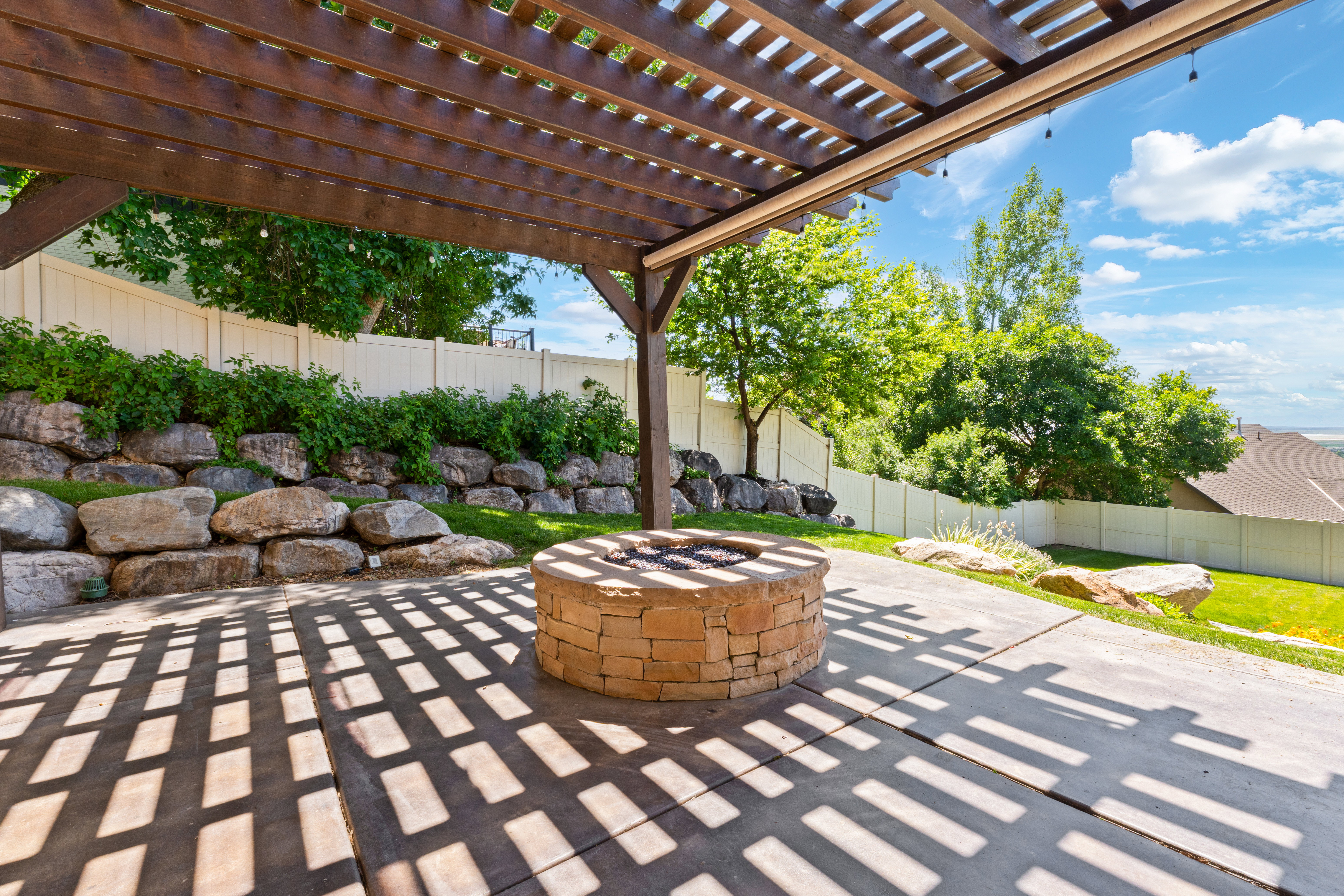 Outdoor patio featuring a wooden pergola, a built-in stone fire pit, concrete paving, a rock retaining wall, and a privacy fence