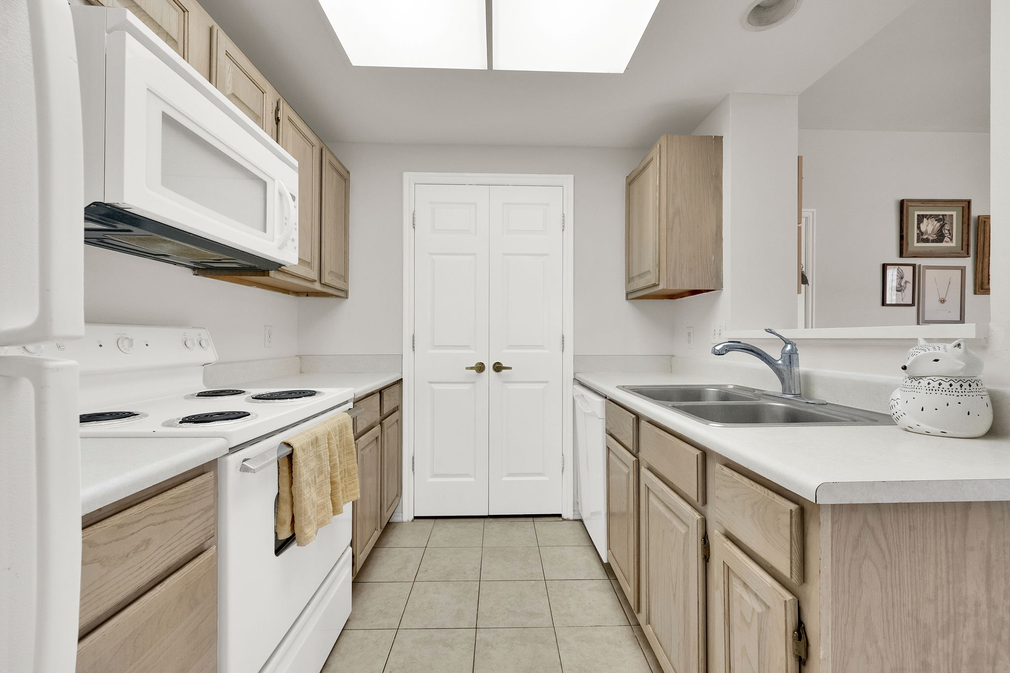 Galley kitchen featuring light wood-finish cabinetry, white appliances, and a dual basin stainless steel sink