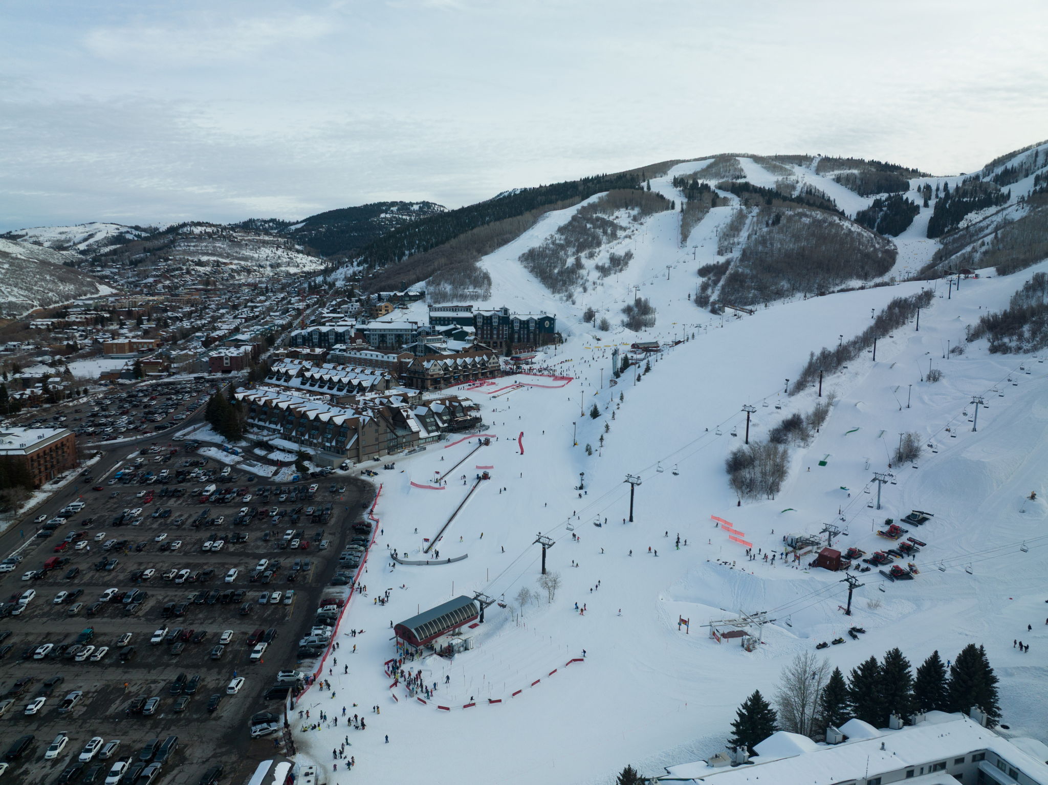 Aerial perspective showcasing a ski resort village nestled at the base of snow-covered mountains, featuring multiple ski runs, chairlifts, and a large parking area