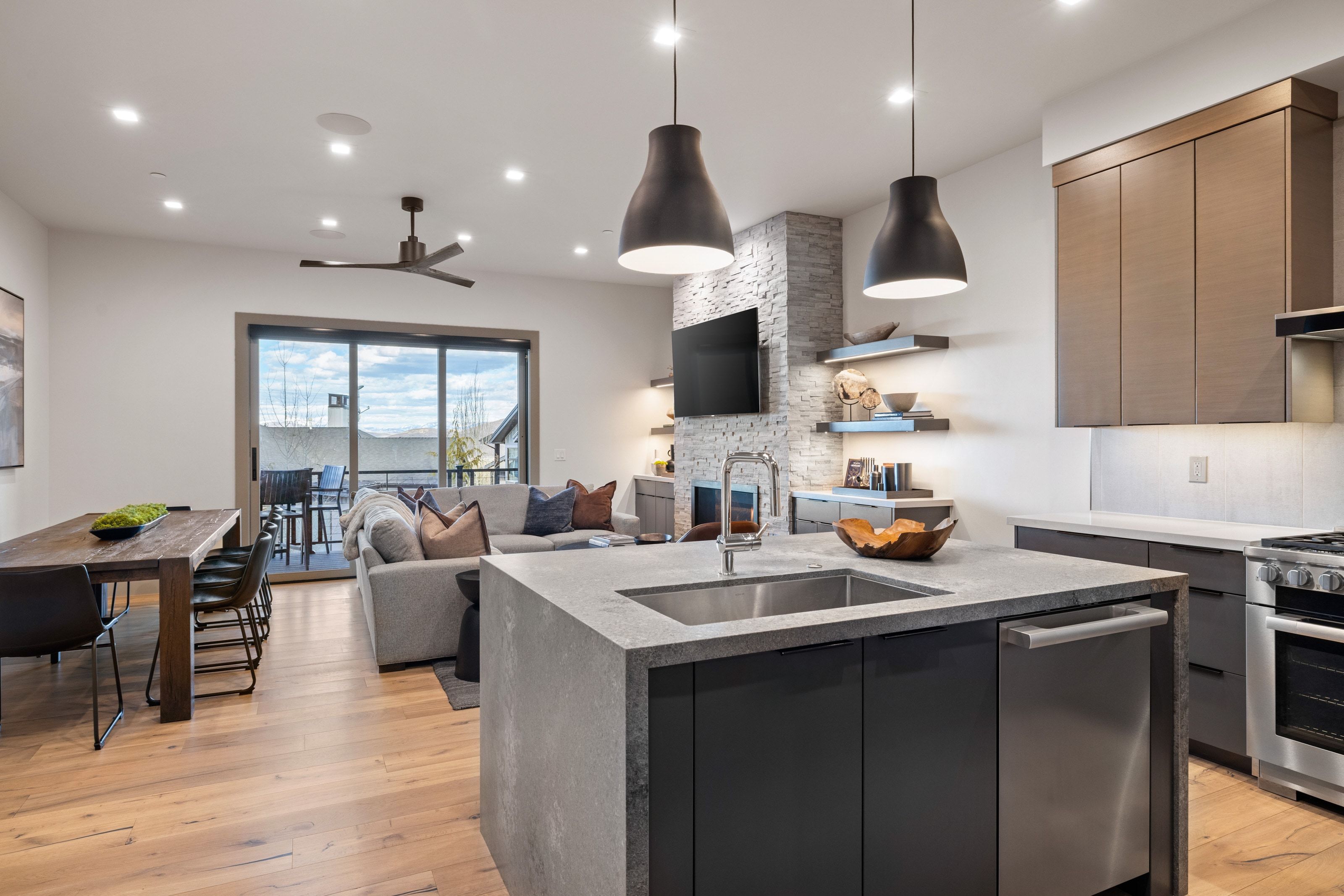 Open-concept living area featuring a kitchen island with integrated sink, wood-finish flooring, a stone-accented fireplace, and sliding glass doors opening to an outdoor space