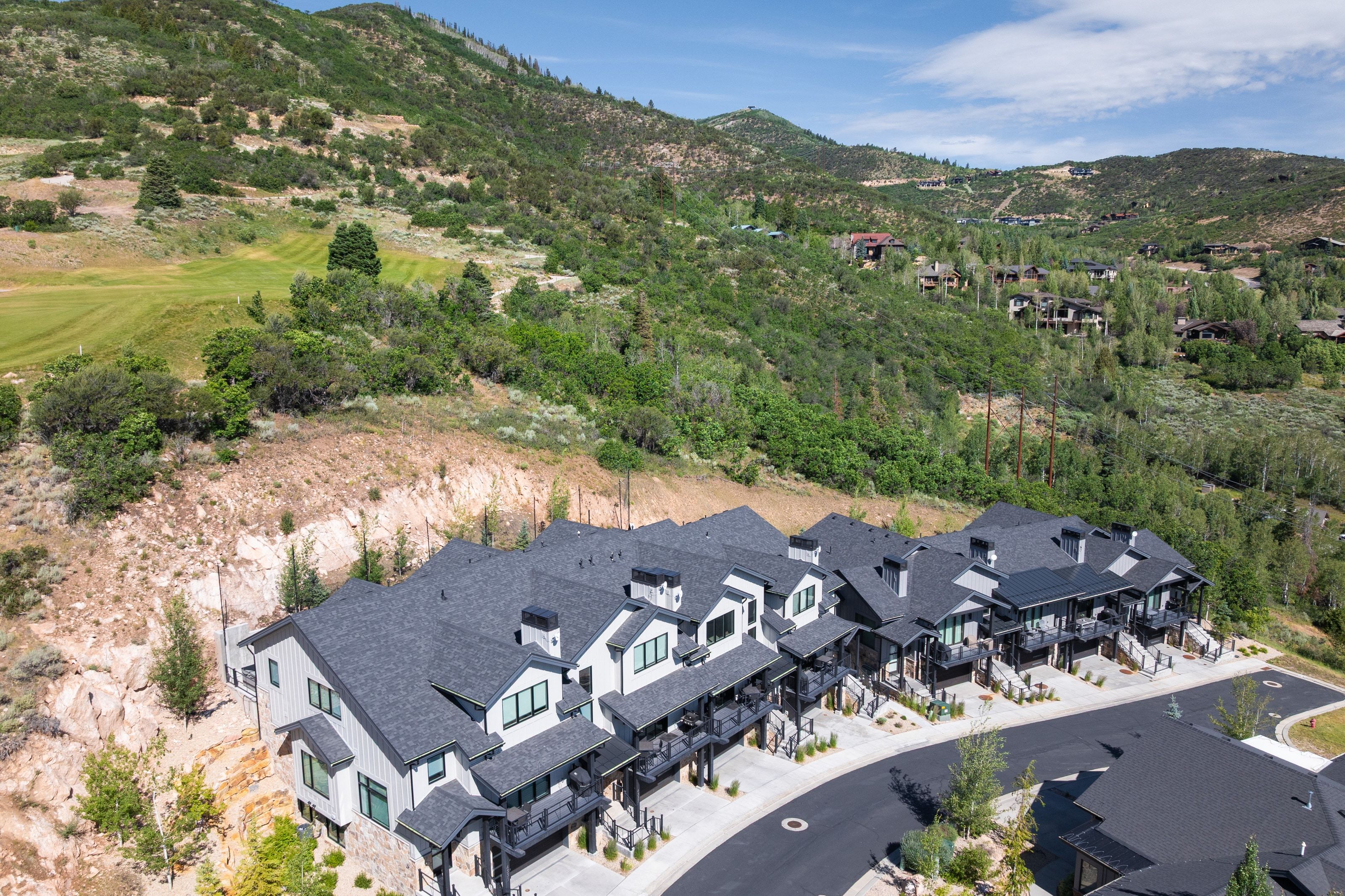 Contemporary townhomes featuring dark gray shingle roofs and light-colored siding, situated against a lush, green mountainside