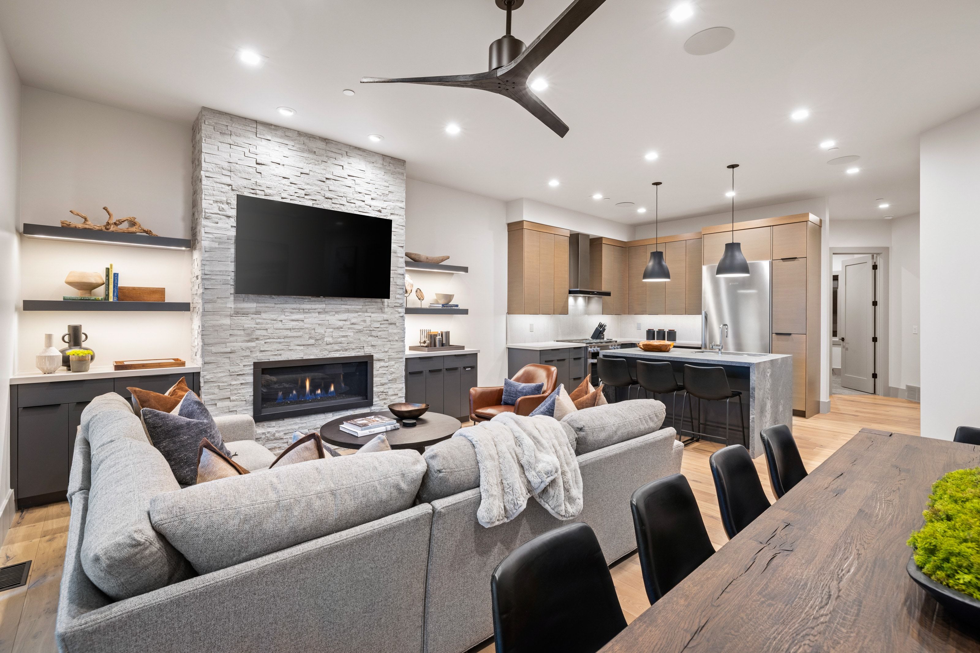 Open-concept living space featuring a stacked stone fireplace, custom built-in shelving with integrated lighting, wood-finish flooring, a kitchen island with seating, and stainless steel appliances