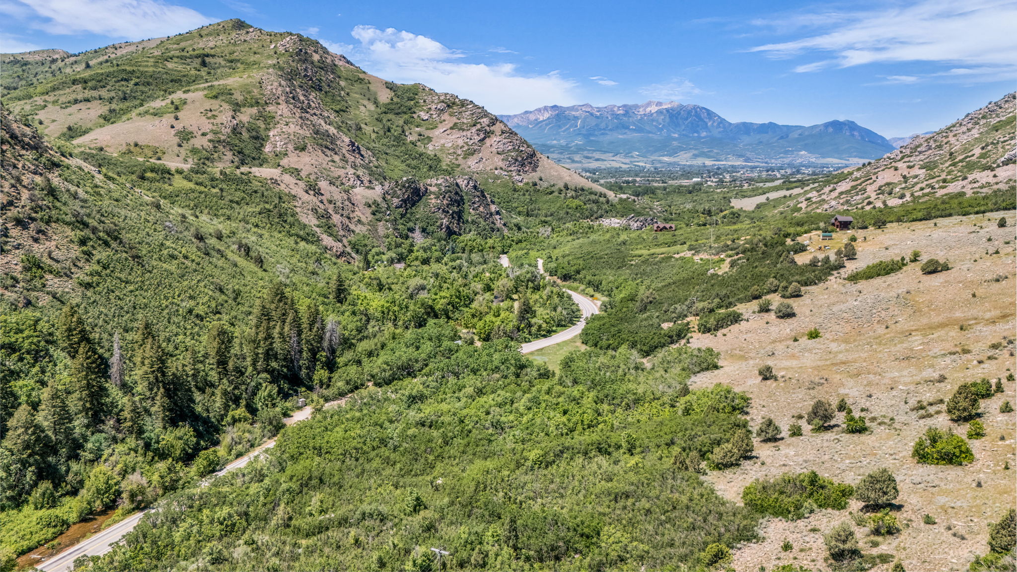 Expansive mountain landscape featuring varied terrain, abundant natural vegetation, a winding road, and distant snow-capped peaks