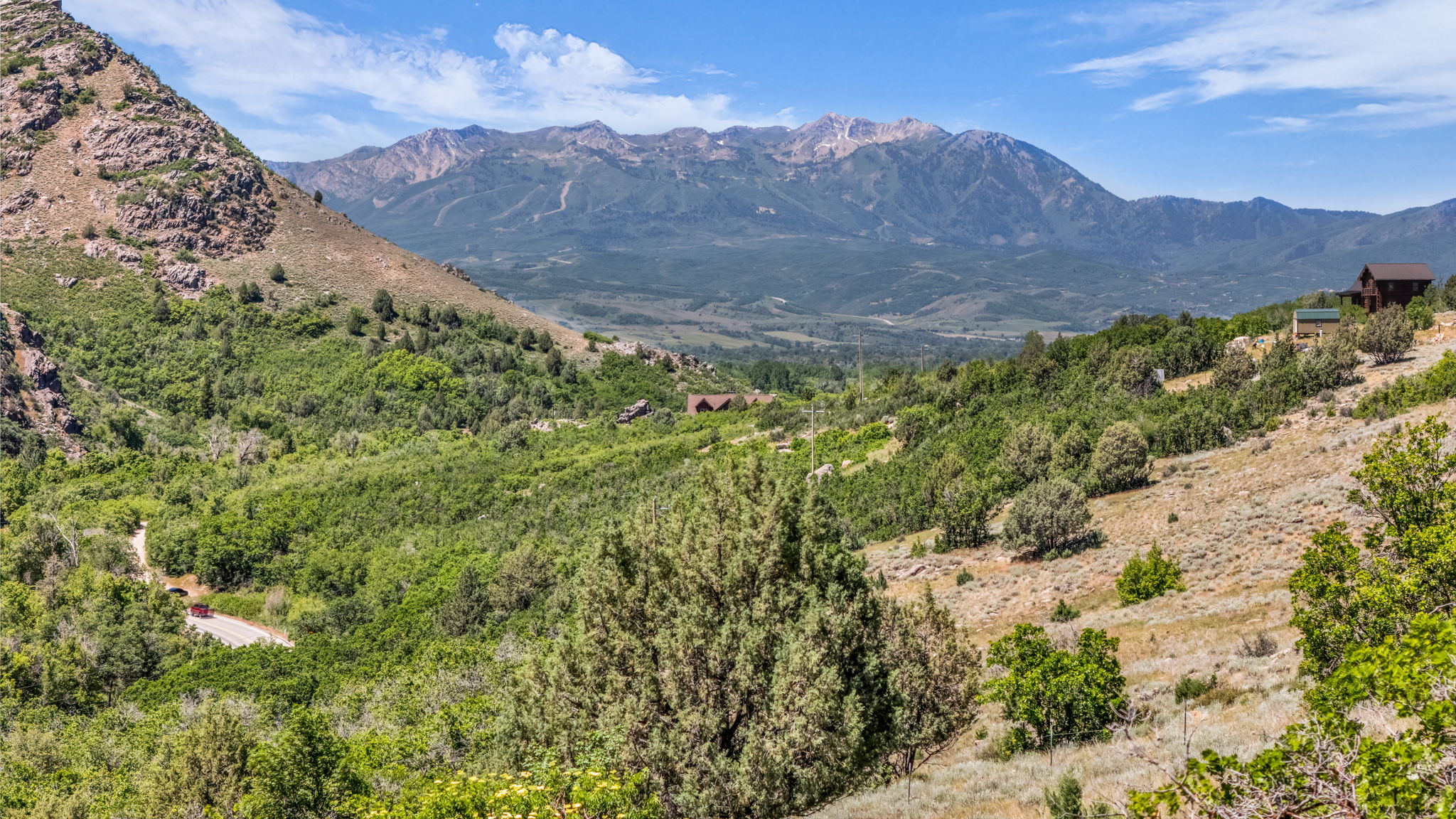 Mountainous landscape with lush green foliage and scattered trees, featuring a distant brown residence with a light-colored roof