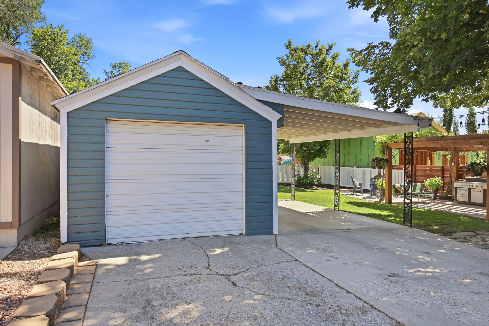 Detached garage featuring blue siding, a white garage door, and a concrete driveway leading to a carport with ornate metal supports
