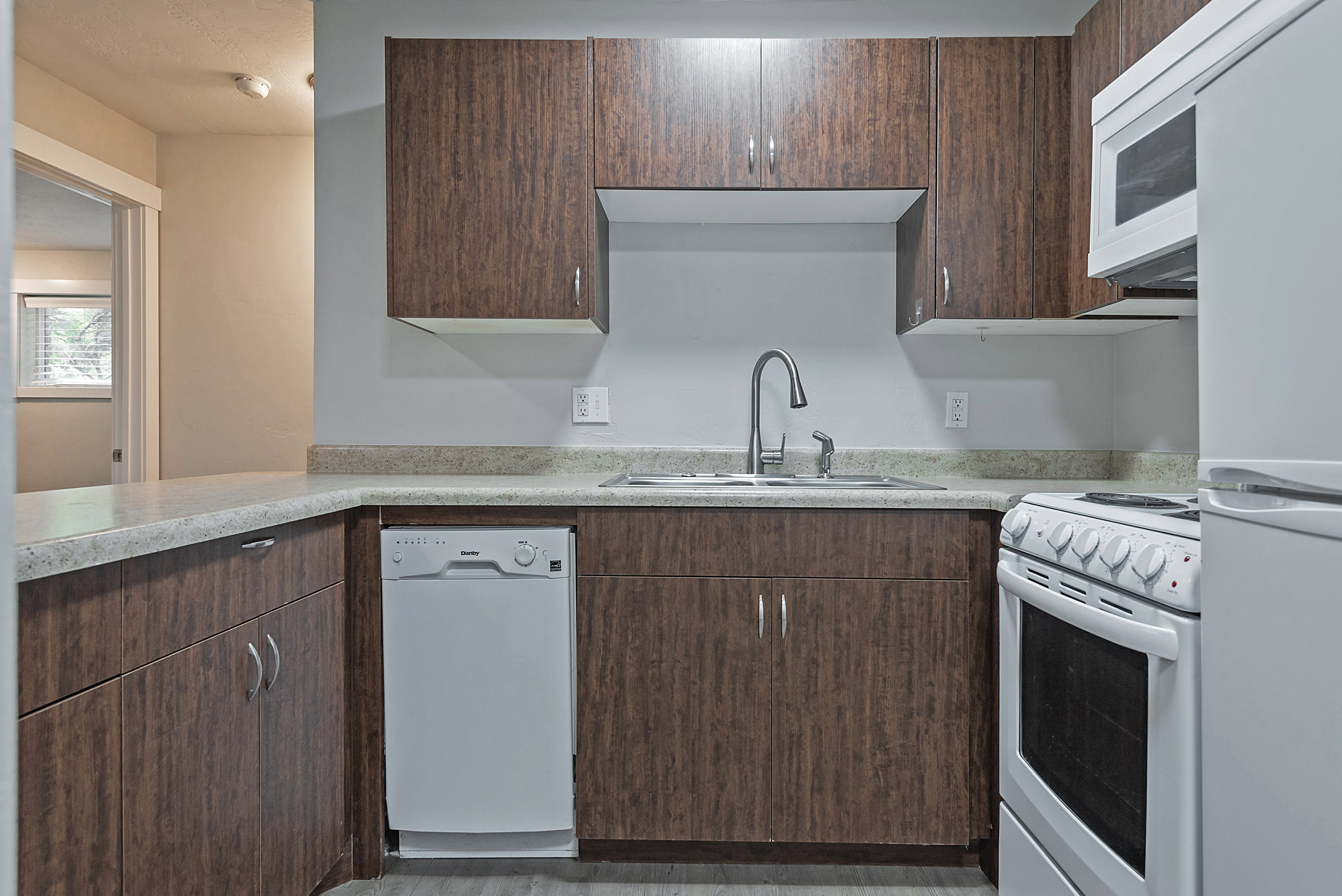 Kitchen featuring wood-finish cabinetry, light-toned countertops, a stainless steel sink with a gooseneck faucet, a white dishwasher, and a white electric range with an overhead microwave