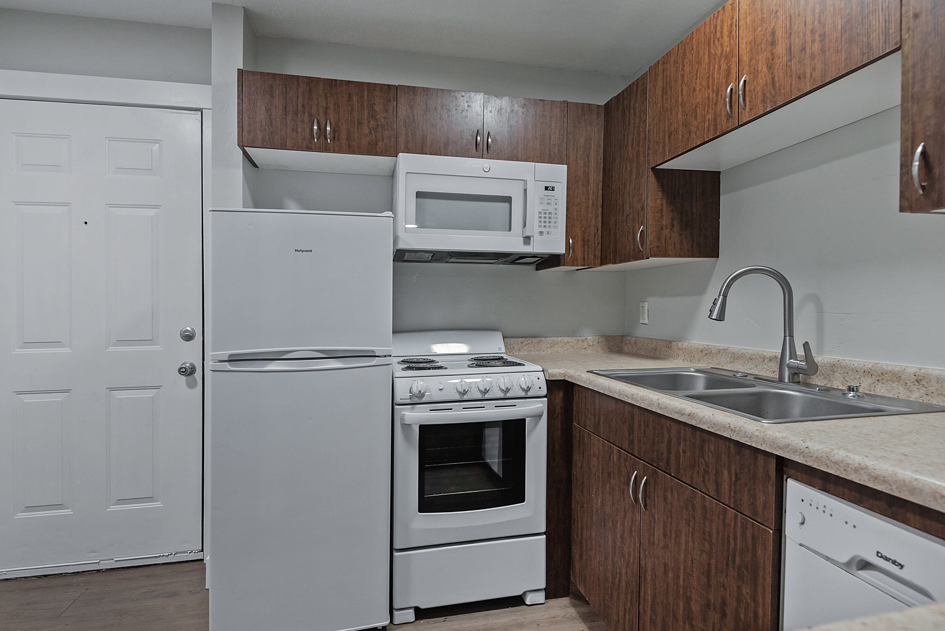 Compact kitchen featuring wood-finish cabinetry, light-toned countertops, and a double basin stainless steel sink with a gooseneck faucet