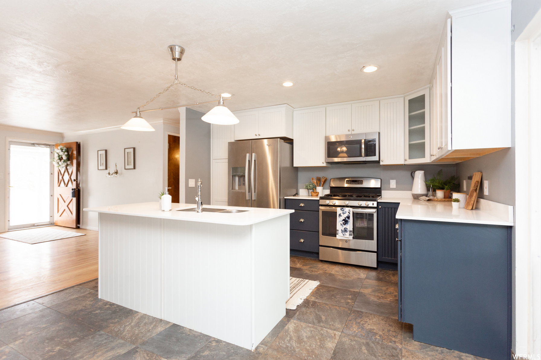 Open-concept kitchen featuring an island with a sink, white cabinetry with glass-front accents, dark lower cabinetry, stainless steel appliances, and stone-finish tile flooring