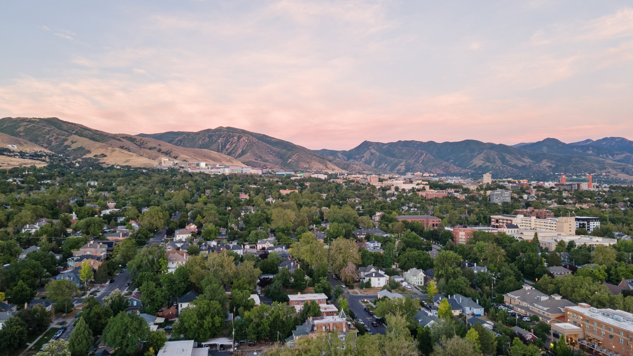 Expansive mountain backdrop framing a dense urban landscape, featuring numerous residential structures, substantial tree canopy, and various commercial buildings