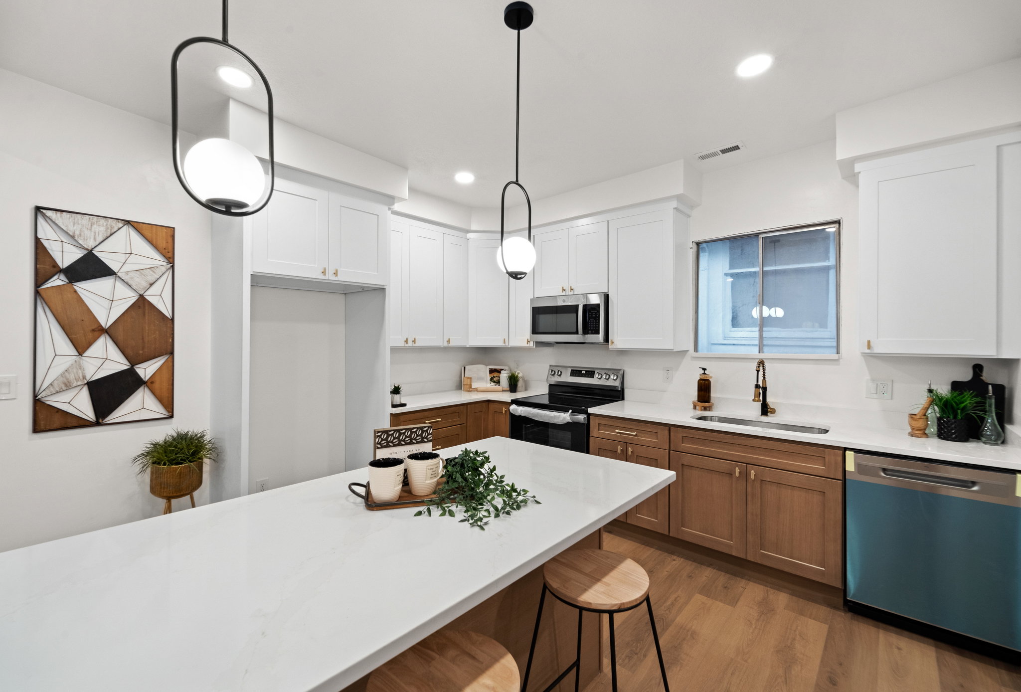Modern kitchen featuring a large central island with a white countertop, two-tone cabinetry with white upper and wood-finish lower units, stainless steel appliances, and wood-finish flooring