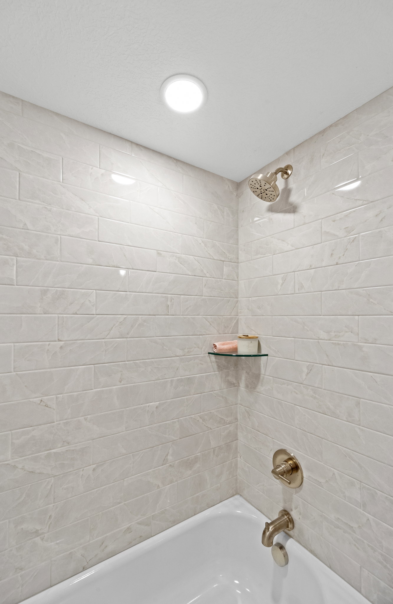 Bathtub and shower combination featuring subway tile surround, recessed lighting, bronze-finish fixtures, and a glass corner shelf