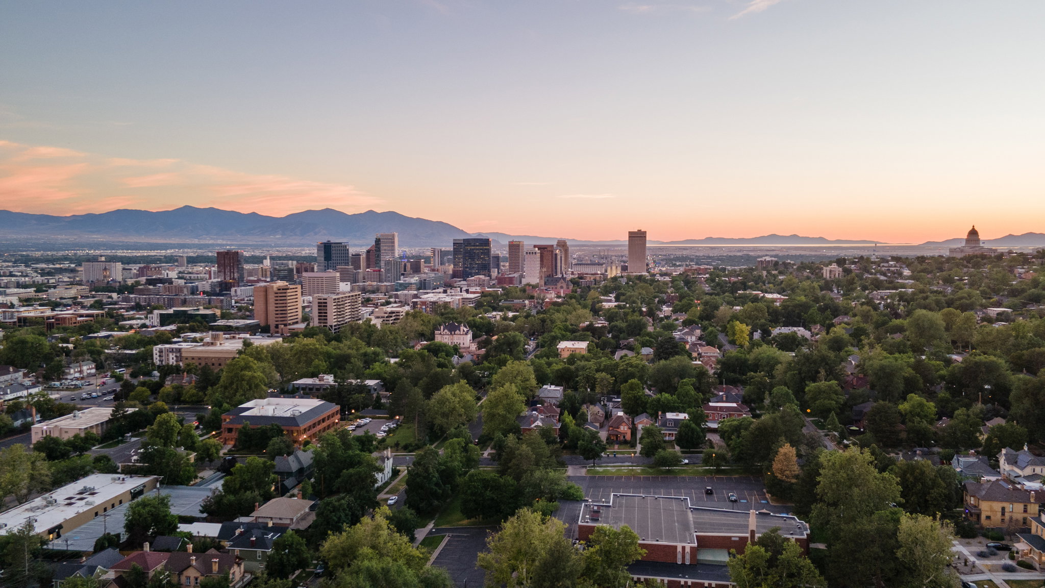 Expansive urban landscape featuring a dense tree canopy, a prominent city skyline with multiple high-rise buildings, and distant mountain ranges under a clear sky