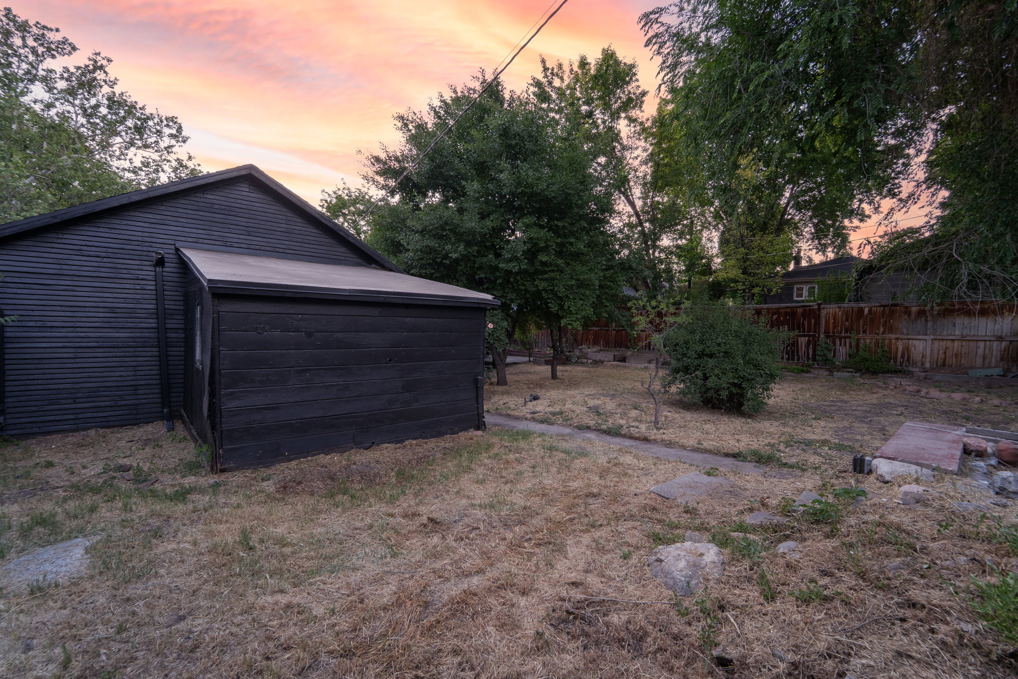 Exterior structure with black horizontal siding and an attached shed, situated on a lot with mature trees and a wood fence perimeter