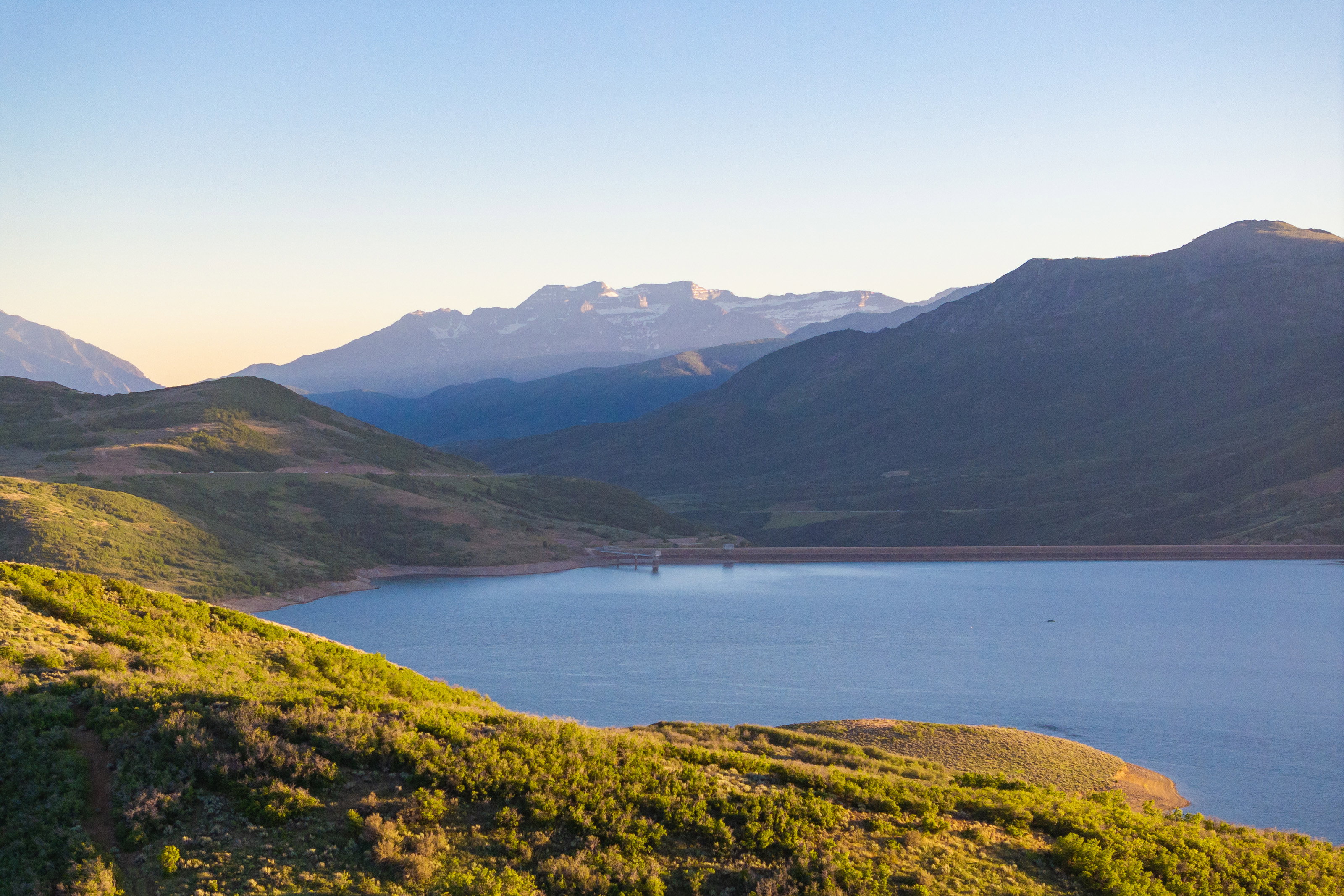 Expansive mountain and lake scenery featuring distant snow-capped peaks, undulating green hills, a large body of water, and a clear horizon