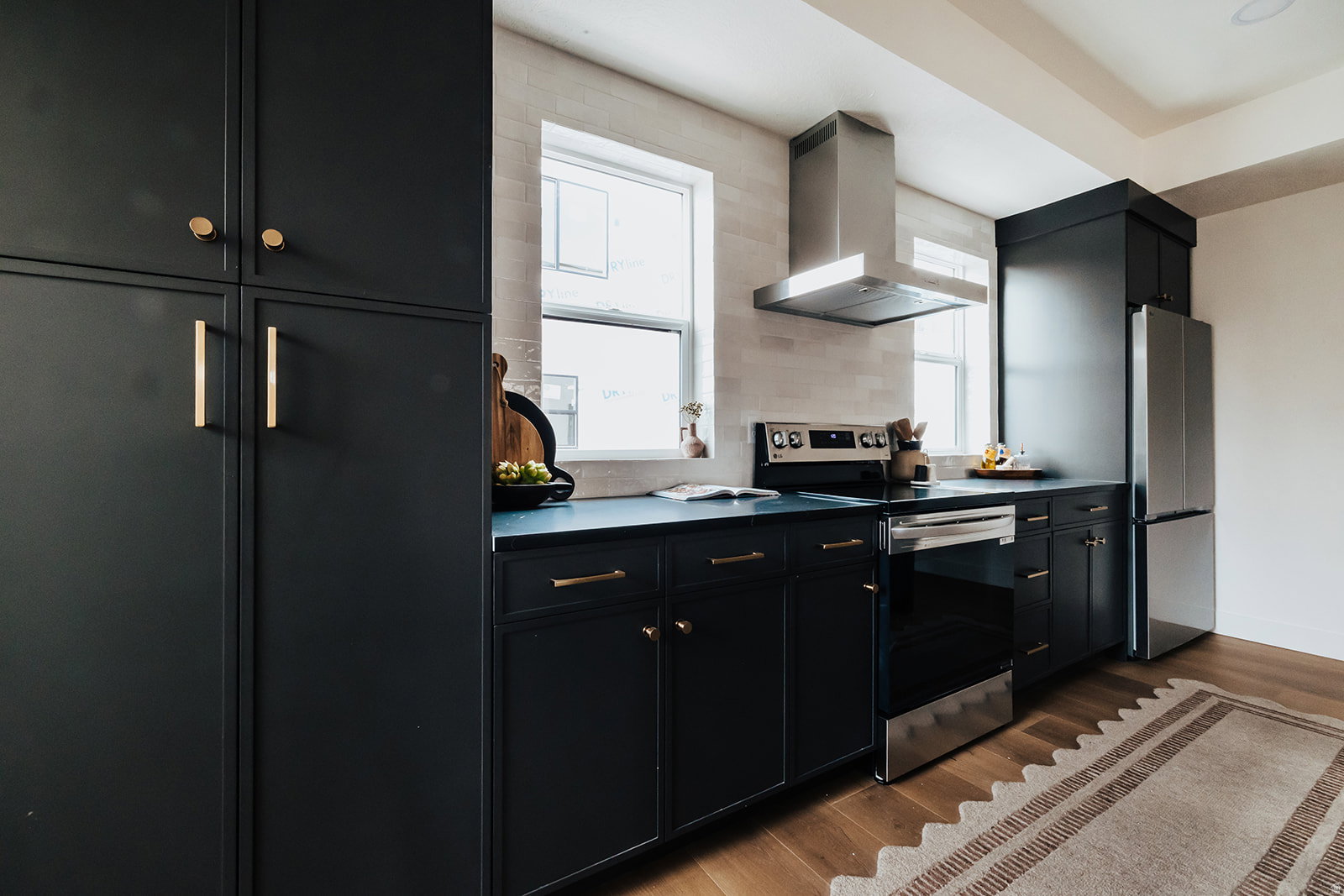 Modern kitchen featuring dark cabinetry with brass hardware, wood-finish flooring, a stainless steel range hood, stainless steel appliances, and a light-toned tile backsplash