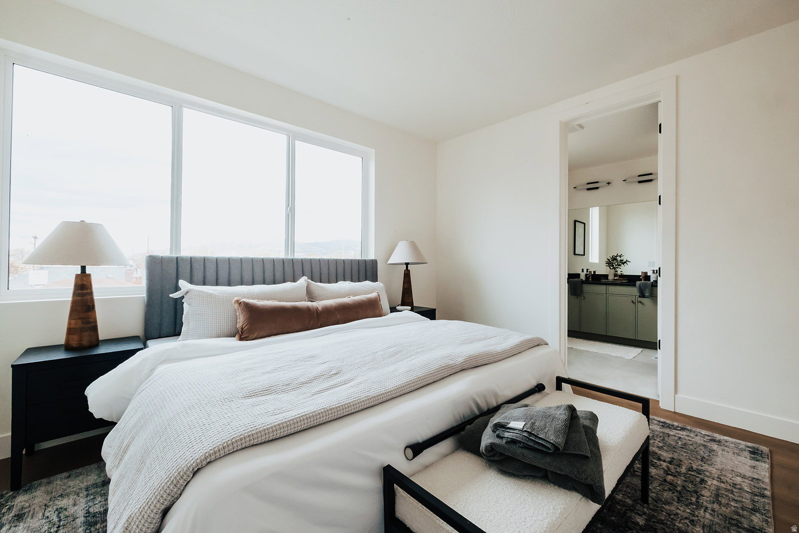 Bedroom featuring large windows, wood-finish flooring, and a doorway opening to a bathroom with green cabinetry and a vanity mirror