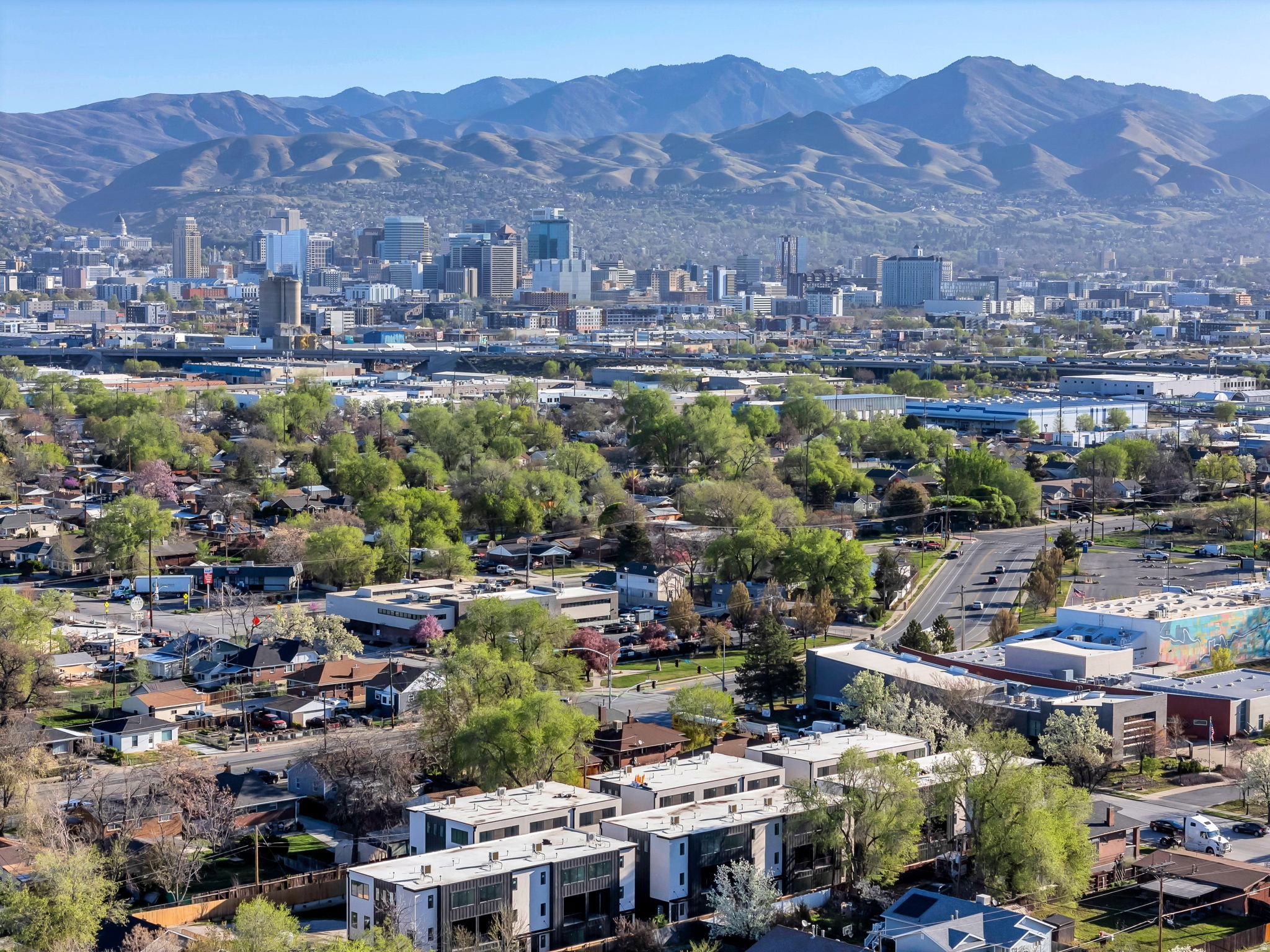Urban skyline featuring a mix of modern and traditional architecture, surrounded by green foliage, with a backdrop of distant mountains