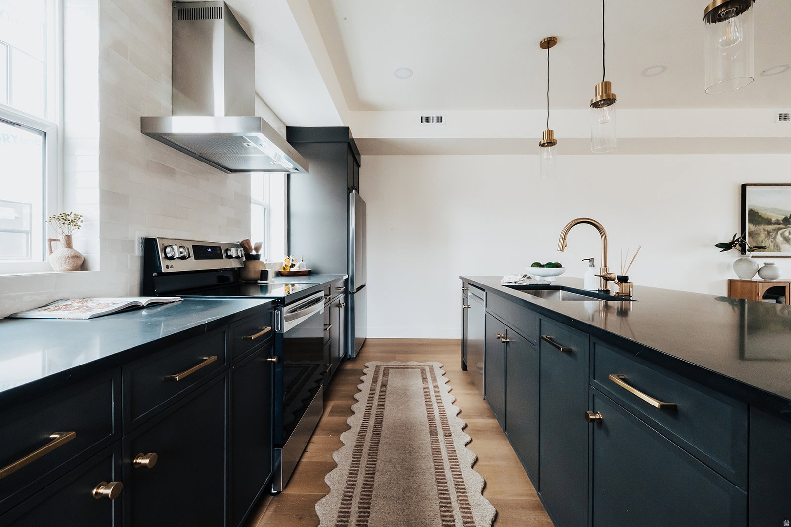 Modern kitchen featuring dark cabinetry with brass hardware, stainless steel appliances, a range hood, and light wood-finish flooring