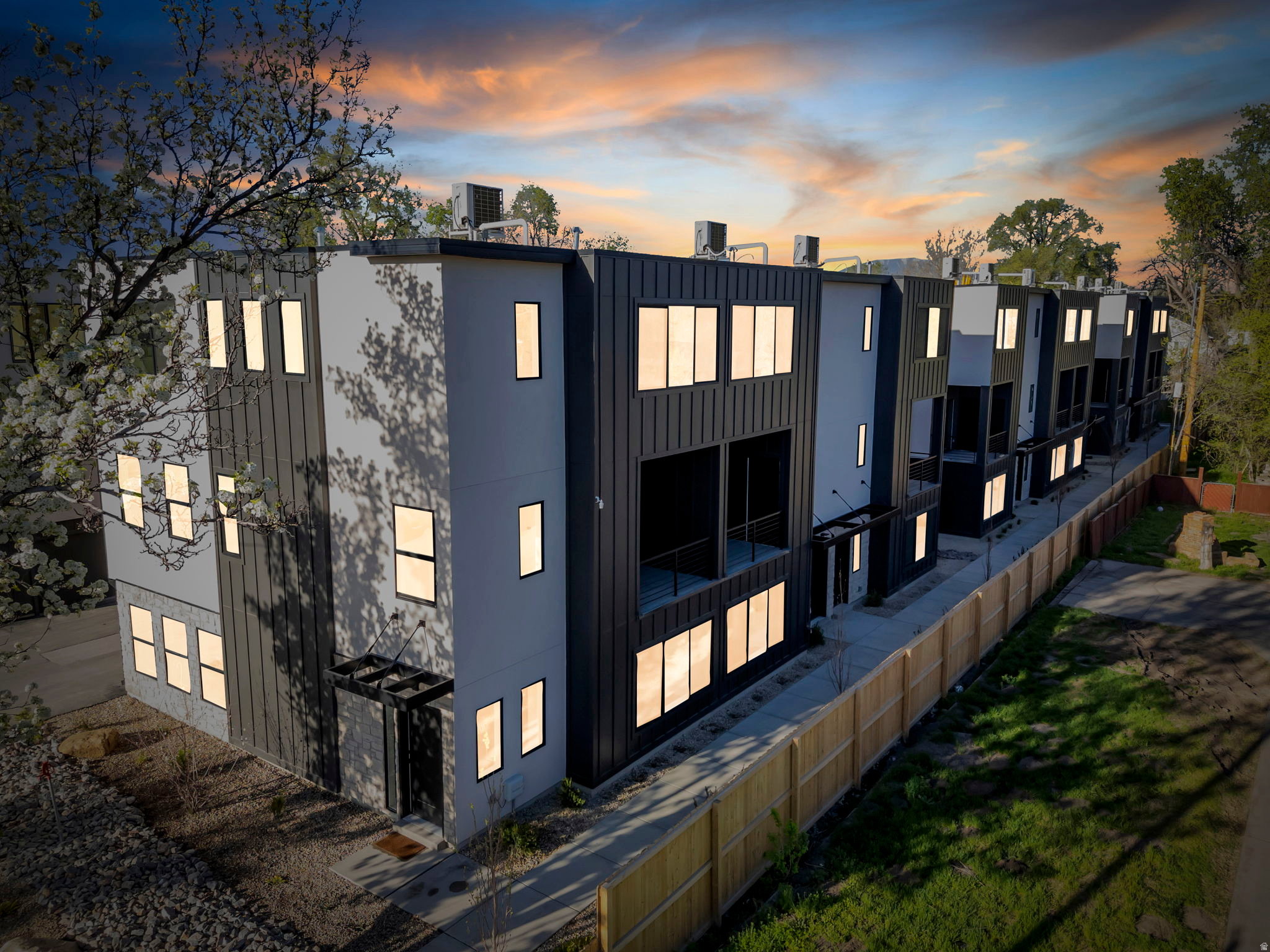 Contemporary townhome-style residences featuring a modern facade with contrasting dark paneling and light-colored siding, multiple windows, private entries, and fenced perimeters