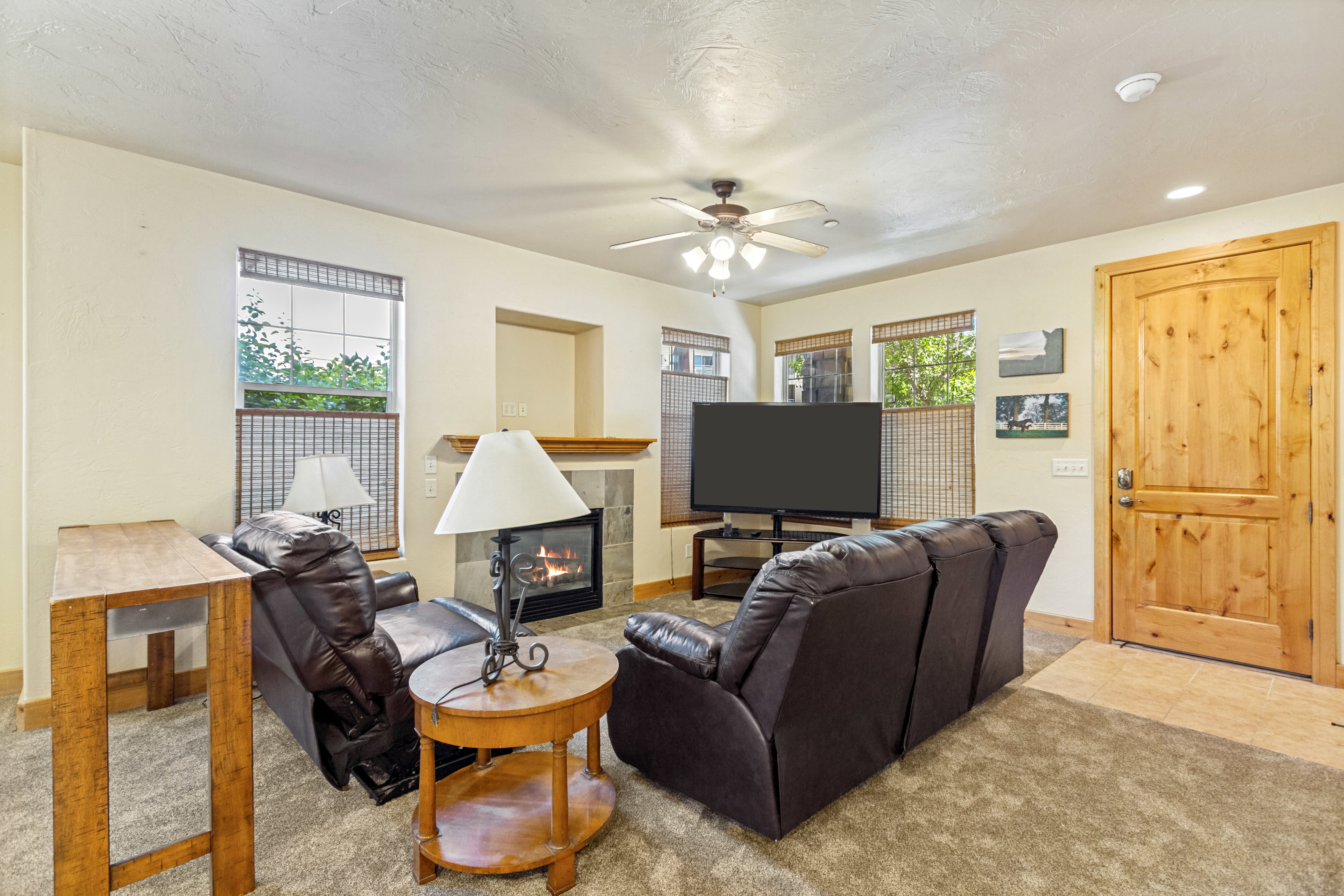 Living area featuring a fireplace with a stone surround and wood mantle, multiple windows with blinds, a ceiling fan, and carpeted flooring