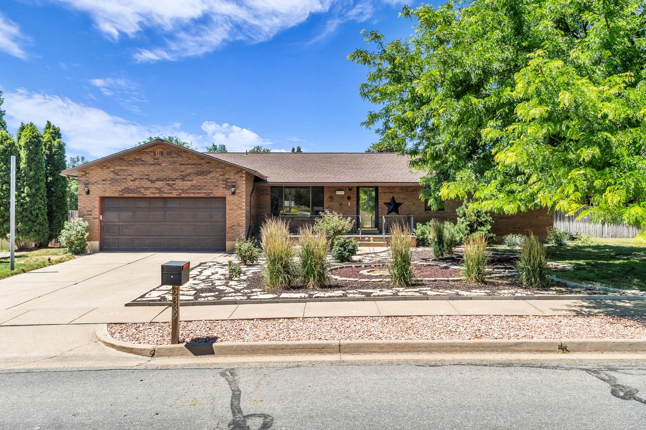 Brick exterior with an attached garage, featuring a paved driveway and mature landscaping with decorative stone beds