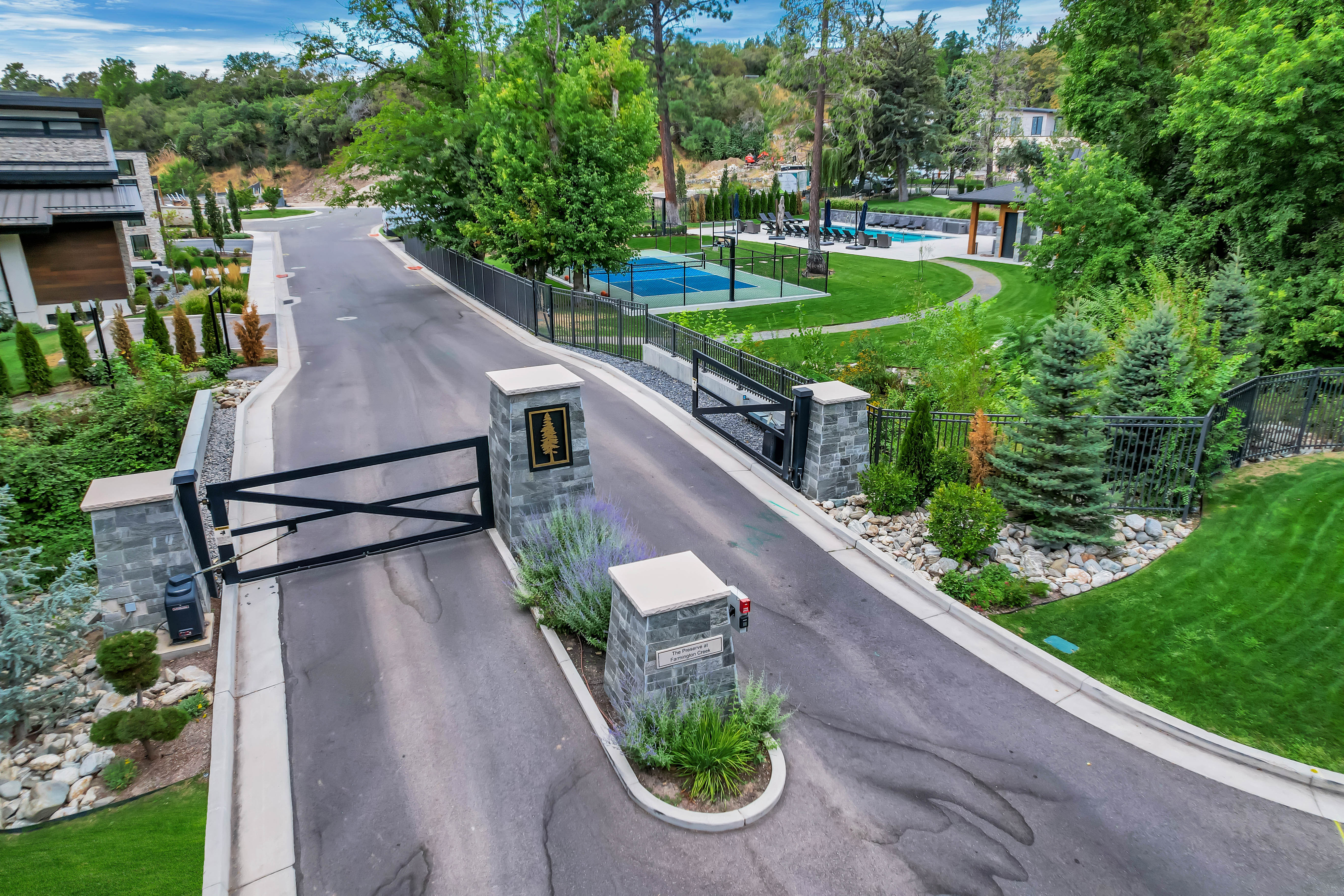Gated community entrance featuring stone pillars, paved drive, mature landscaping, and proximity to community amenities including a pool and sports court