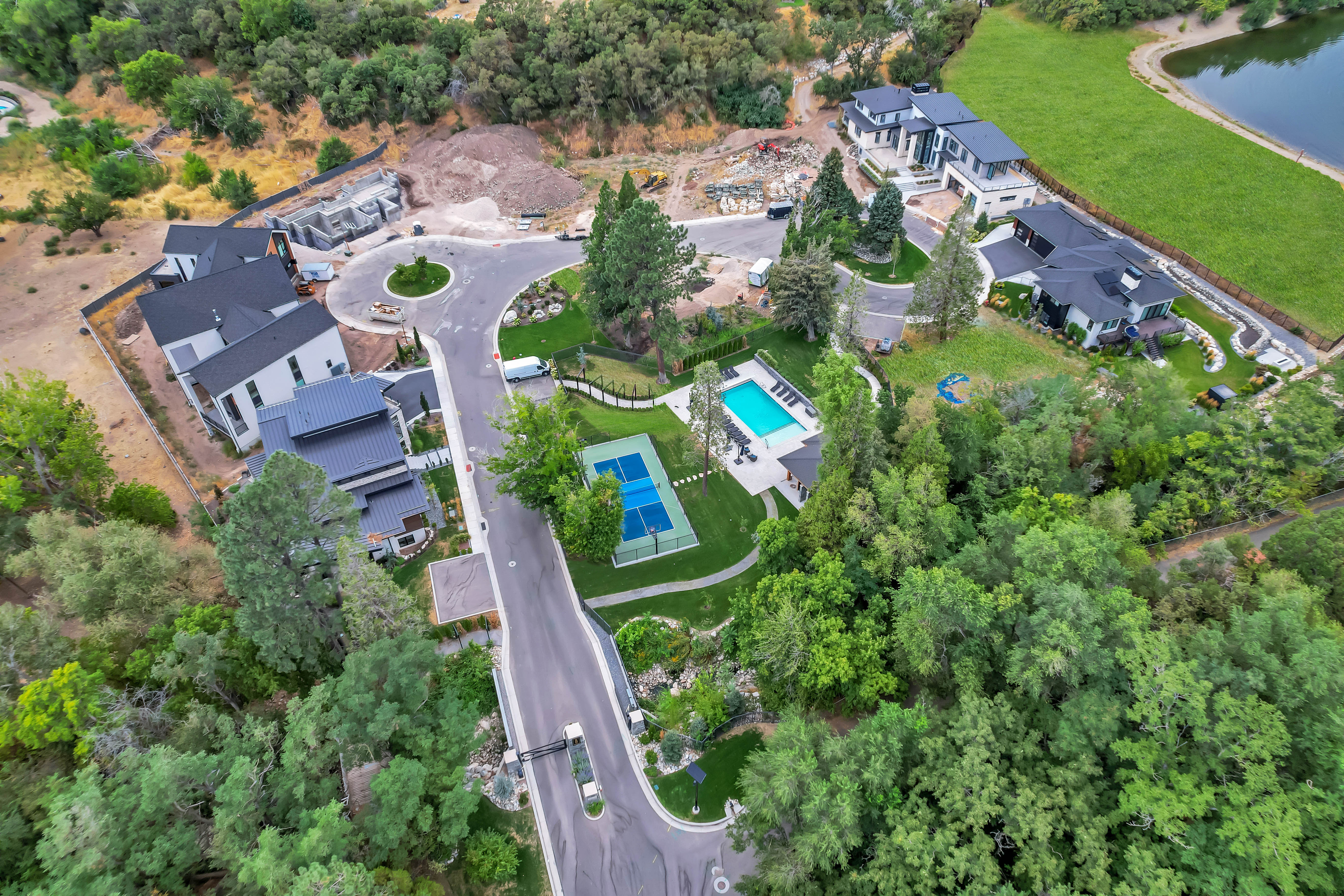 Aerial view of a residential community with contemporary architecture, featuring a private swimming pool, a dedicated tennis court, mature tree landscaping, and a curvilinear paved road