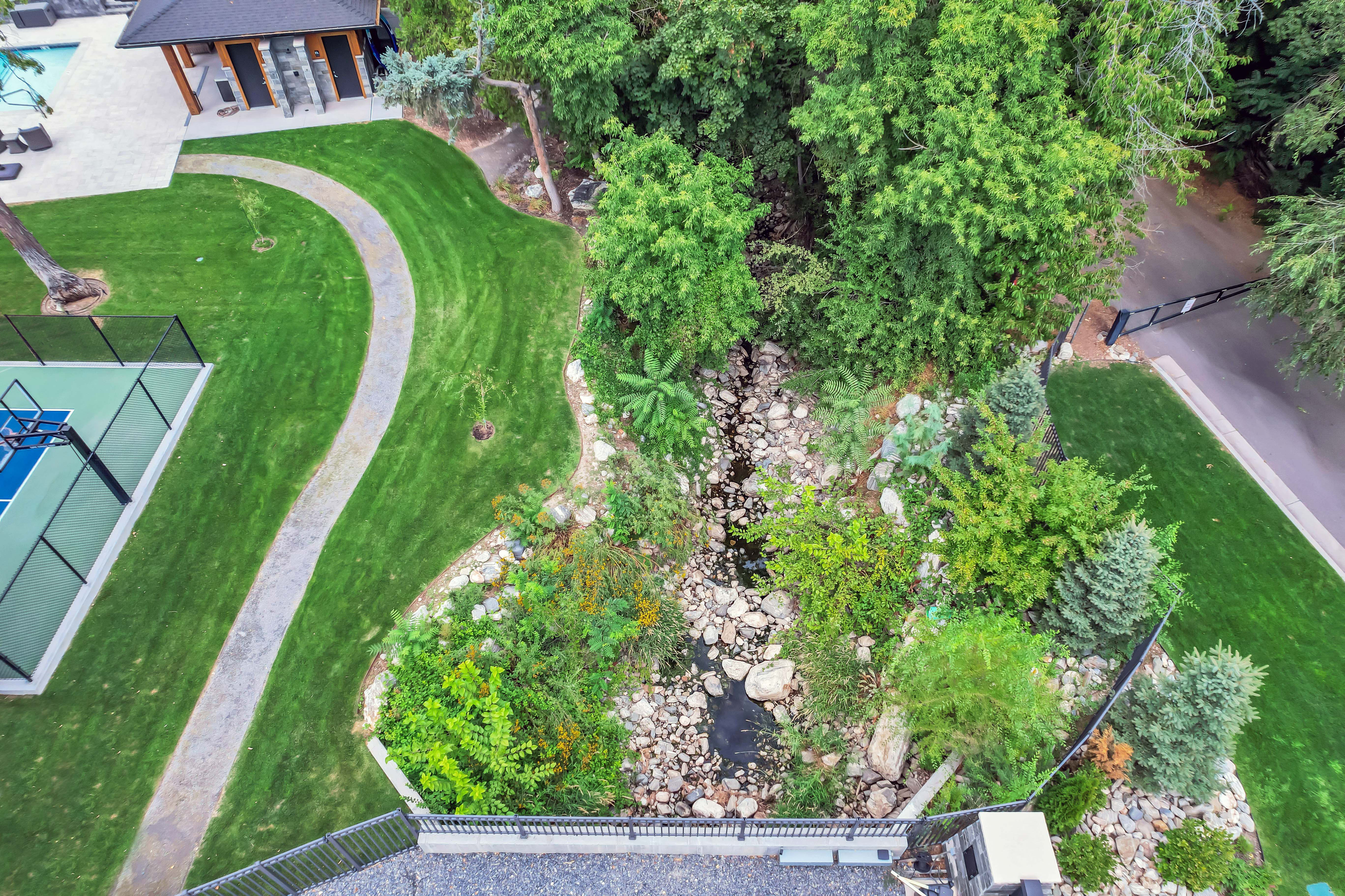 Expansive outdoor recreation area featuring a full-size sports court, a meandering paved pathway, a natural rock creek bed, a stone patio with an adjacent structure, and mature landscaping
