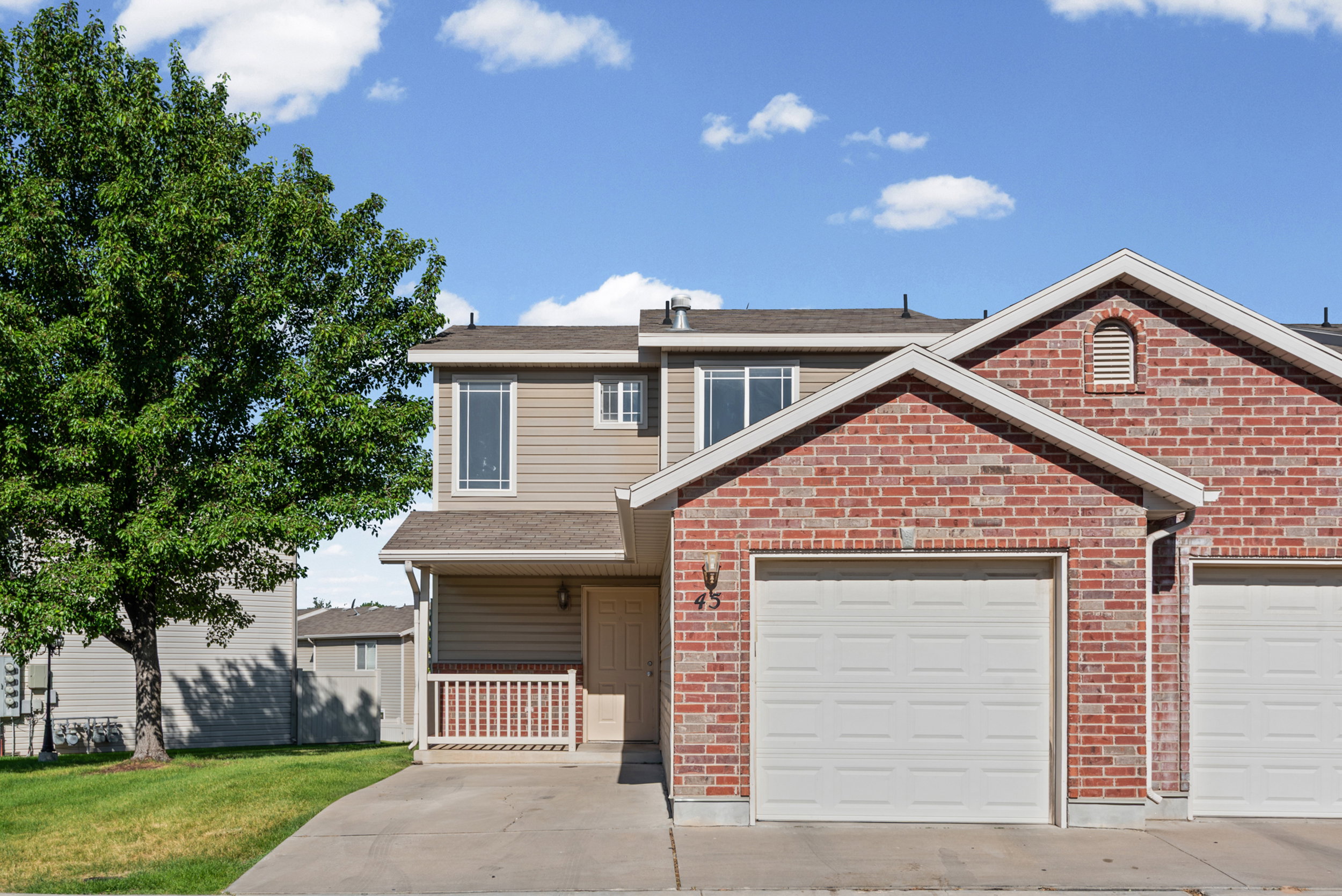 Brick and siding exterior featuring an attached garage, covered front porch with white railing, and a mature tree