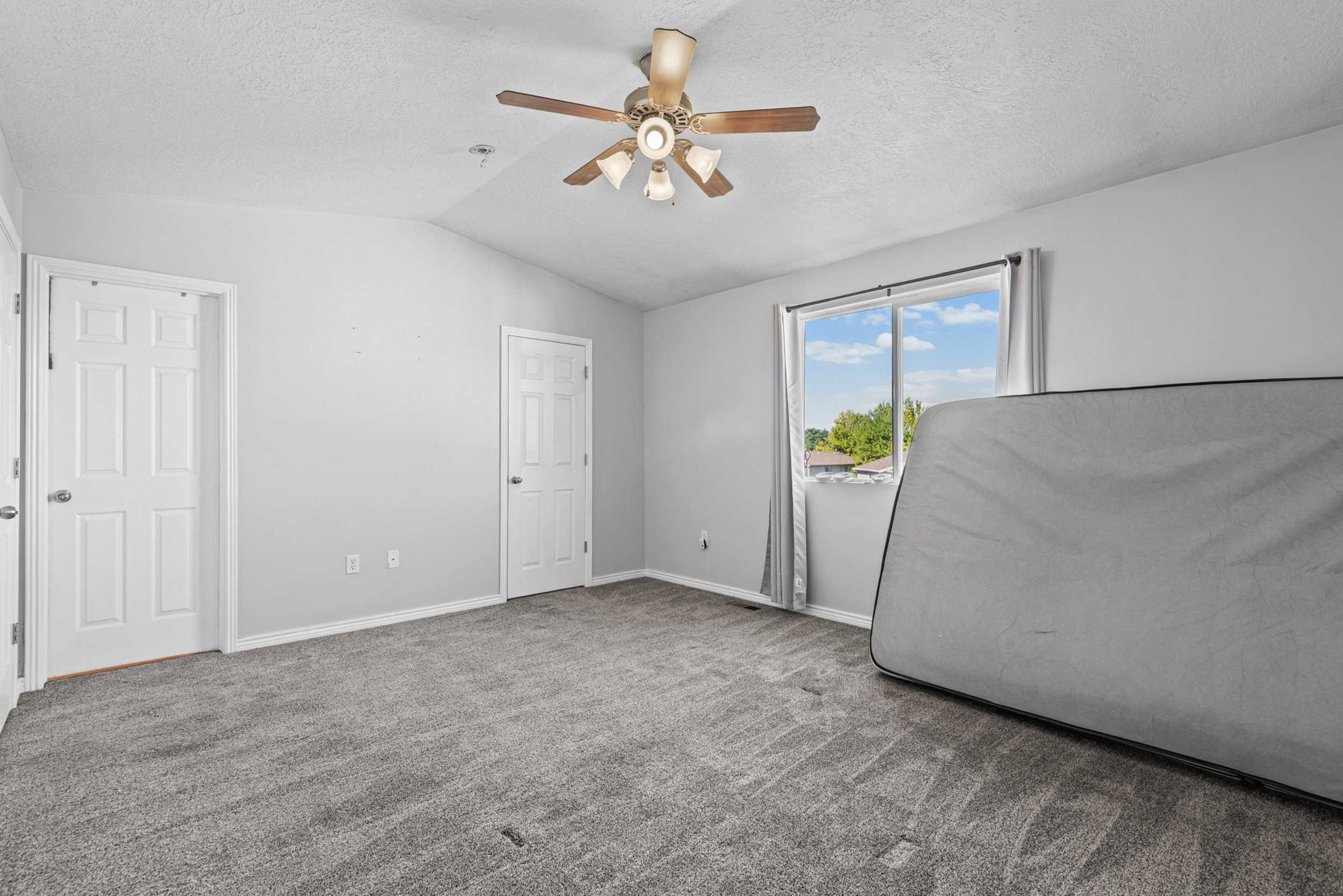 Vaulted ceiling room featuring light gray carpeting, twin pane windows with gray curtains, and a ceiling fan with light fixture