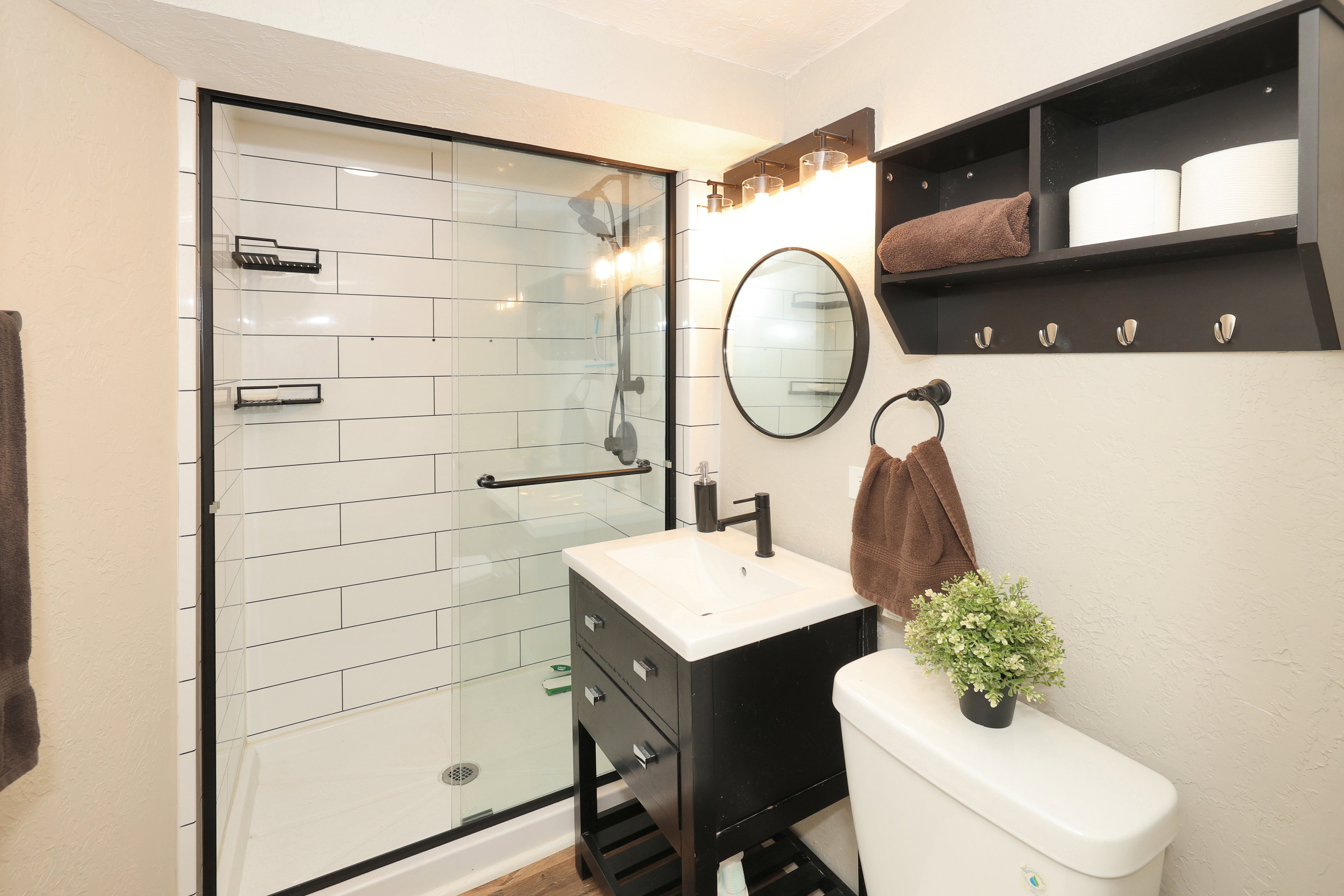 Bathroom featuring a glass-enclosed shower with subway tile surround, a black vanity with a white integrated sink, a circular mirror, a wall-mounted light fixture, and a built-in shelving unit with hooks