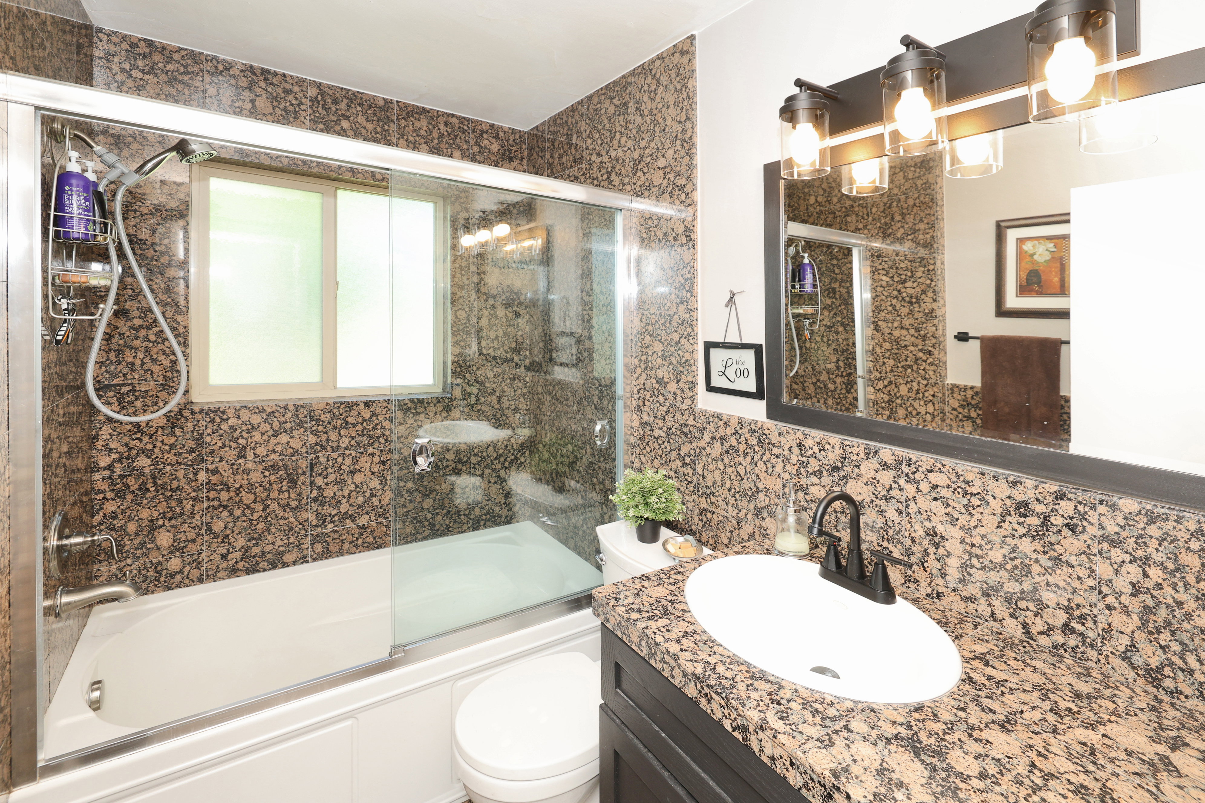 Bathroom featuring a shower-tub combination with sliding glass doors, a vanity with a white oval sink, a black faucet, and a granite-patterned countertop