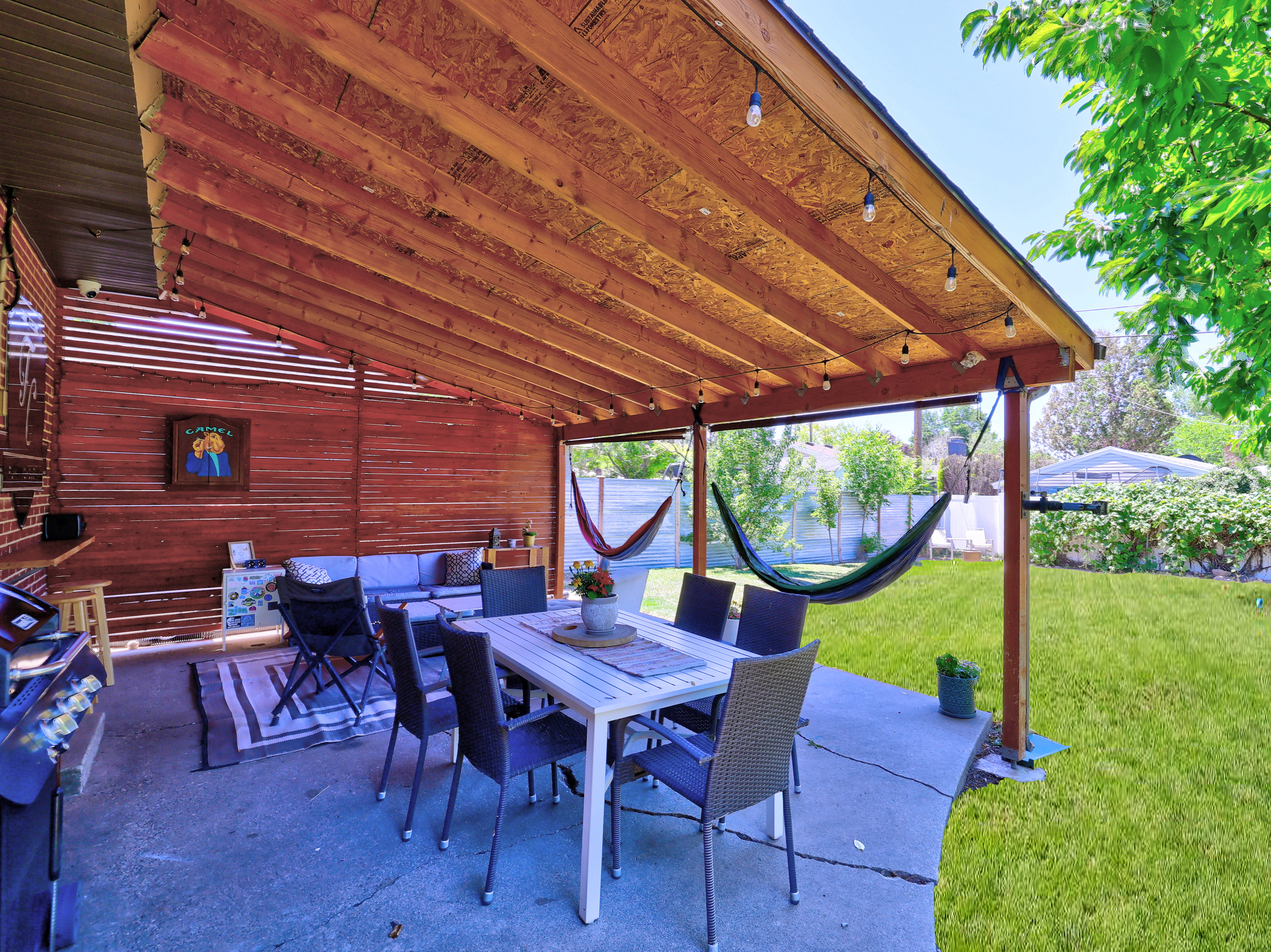 Expansive covered patio featuring a wood-beam ceiling with string lighting, concrete flooring, a partial wood-slat wall, and direct access to a manicured lawn