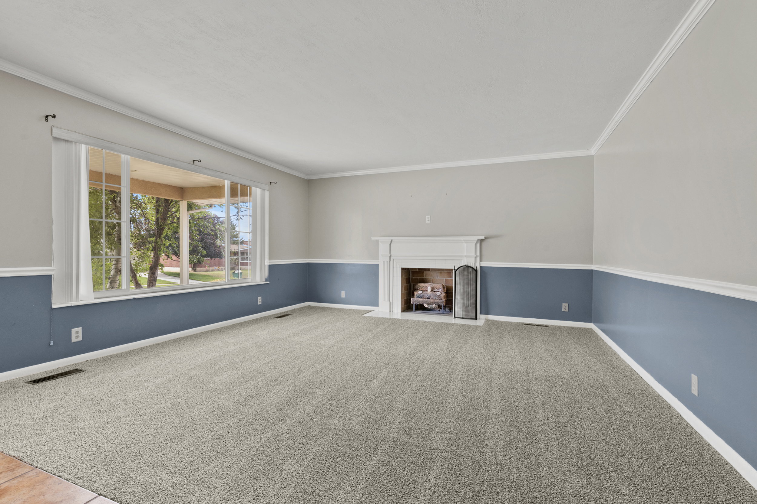 Spacious carpeted room featuring a decorative fireplace with a white mantel, large bay window, two-tone wall paint with a chair rail, and crown molding