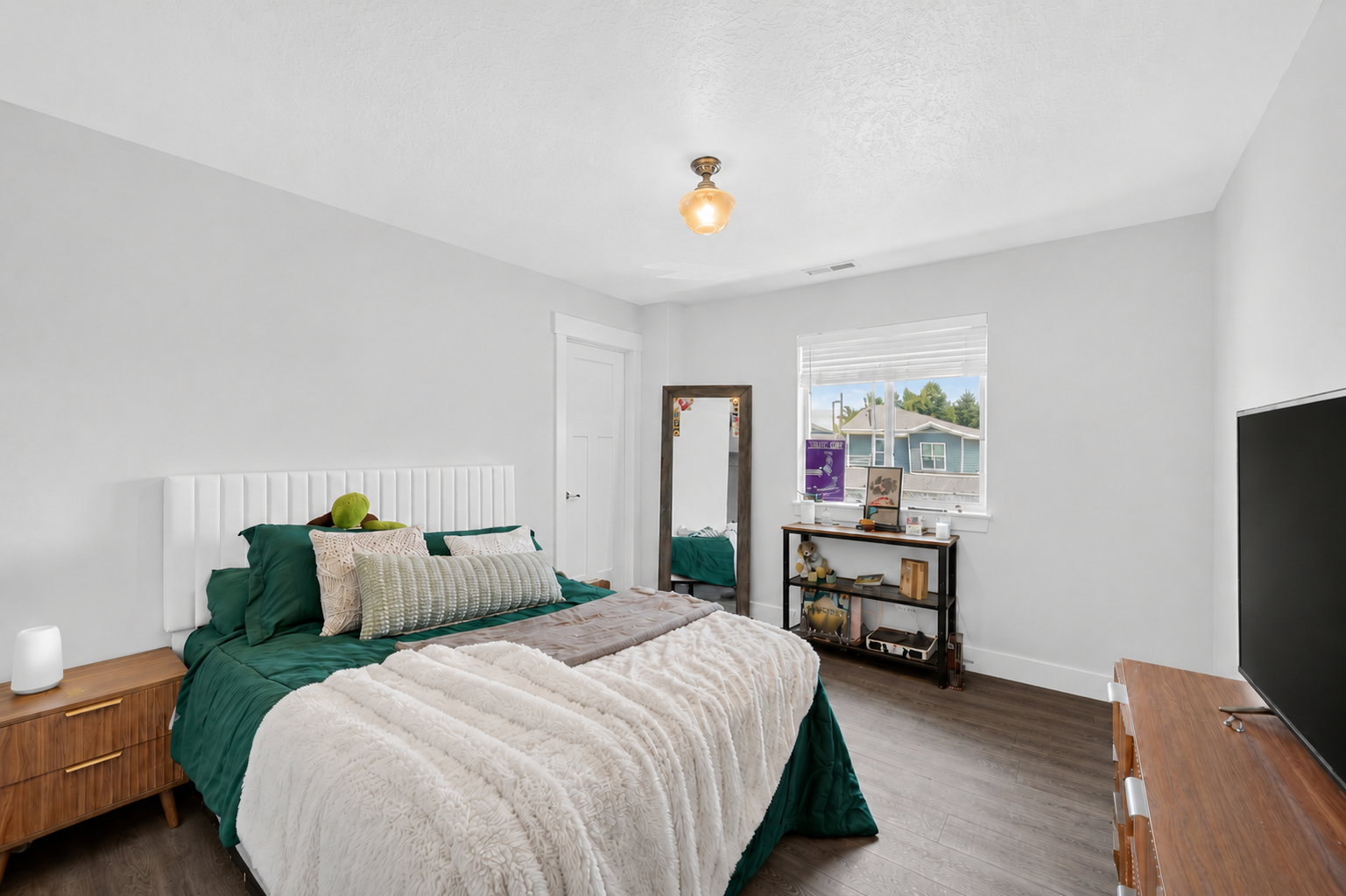 Bedroom featuring wood-finish flooring, a contemporary white paneled headboard, a white painted door with dark hardware, a single window with blinds, and a built-in ceiling light fixture